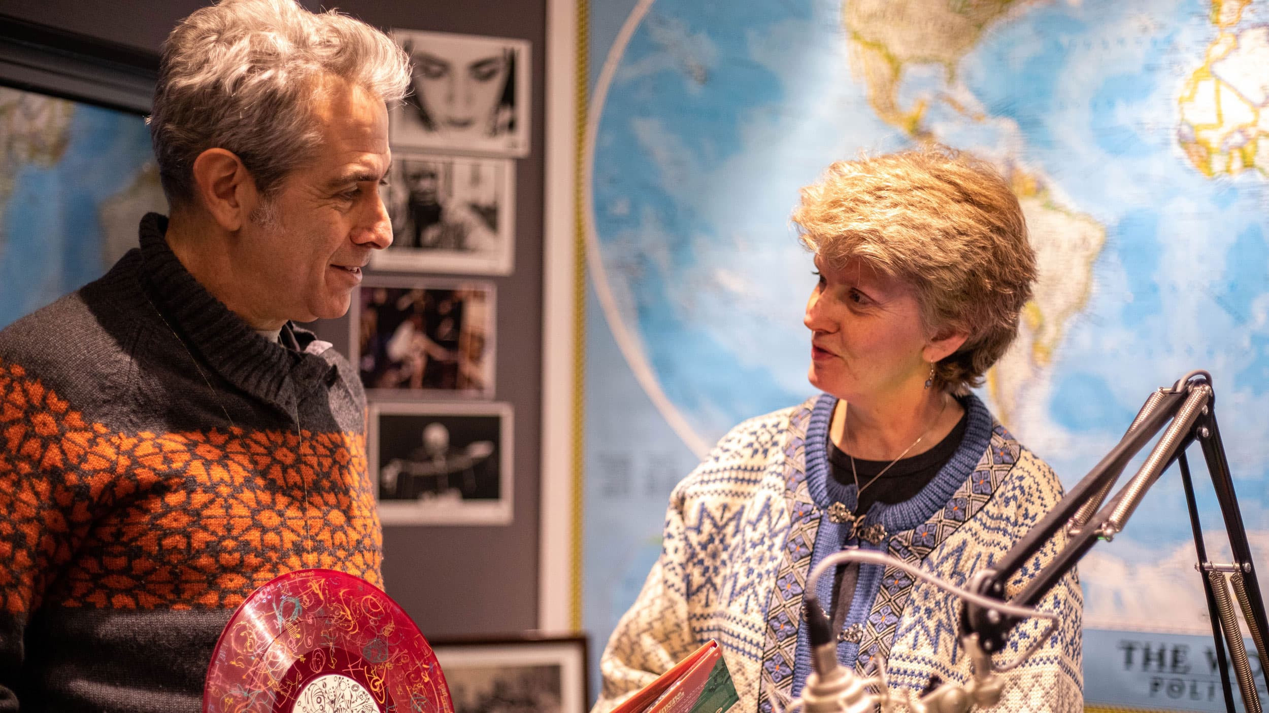 Marco Werman is shown standing holding a red record across from April Peavey with a microphone stand inbetween them