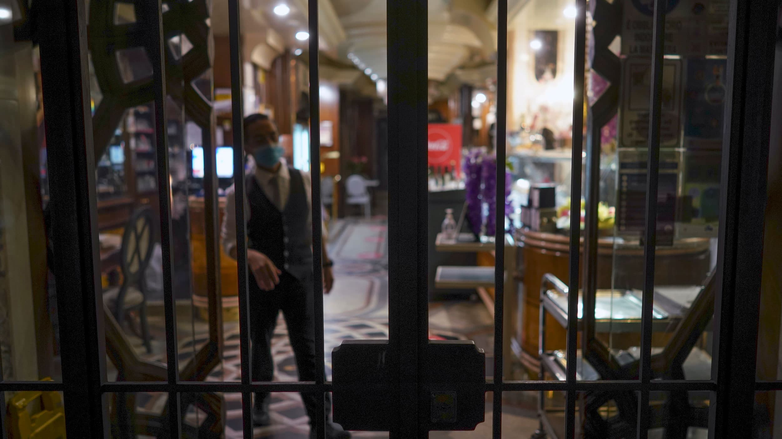 A man is shown wearing a face mask and tailored vest behind the closed-gated doors of a store front.