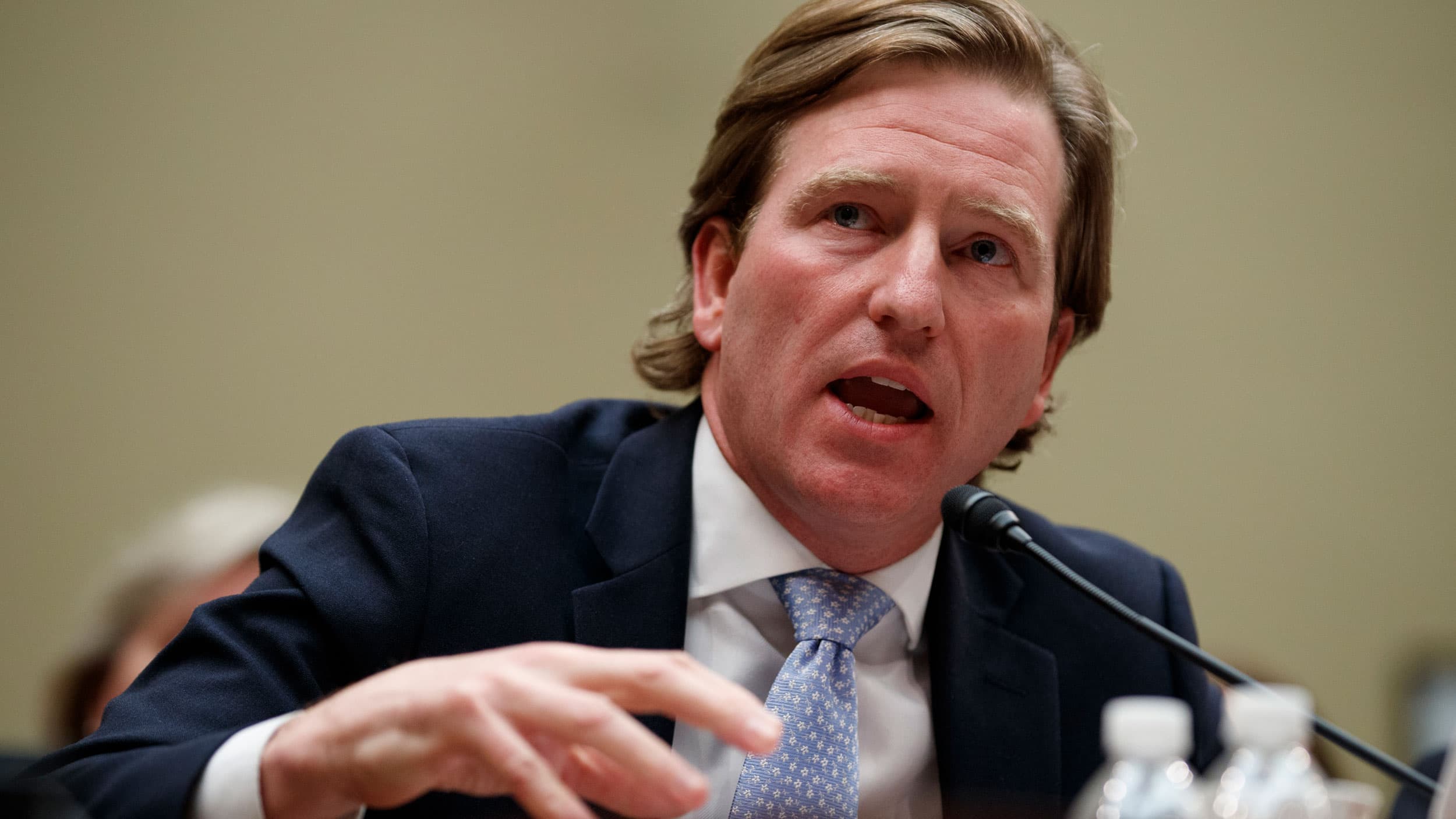 Christopher Krebs is shown seated and wearing a blue suit and light blue tie while speaking into a small microphone.