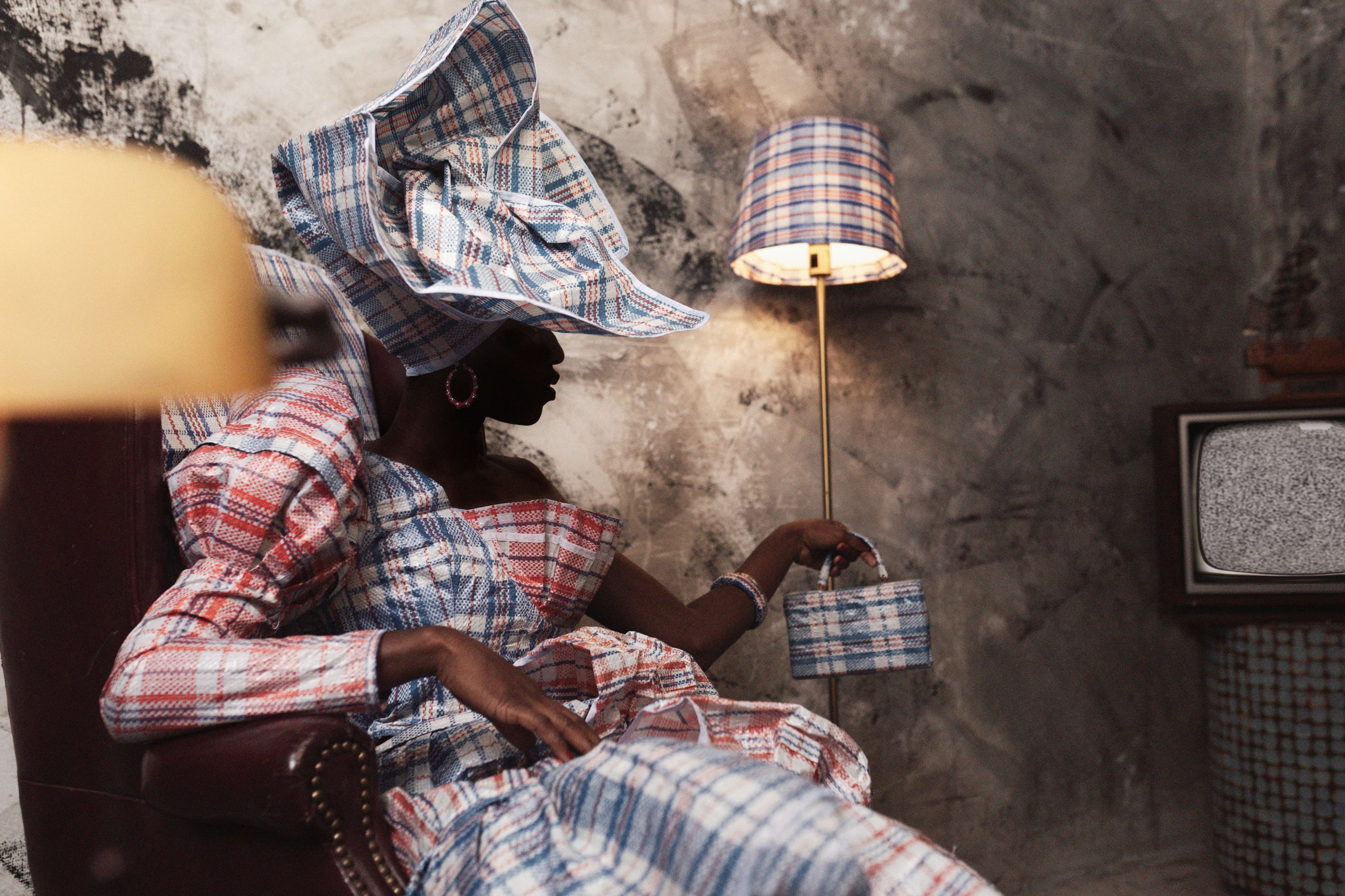 A woman wearing a plaid head wrap and dress sits near a lamp with same design on shade