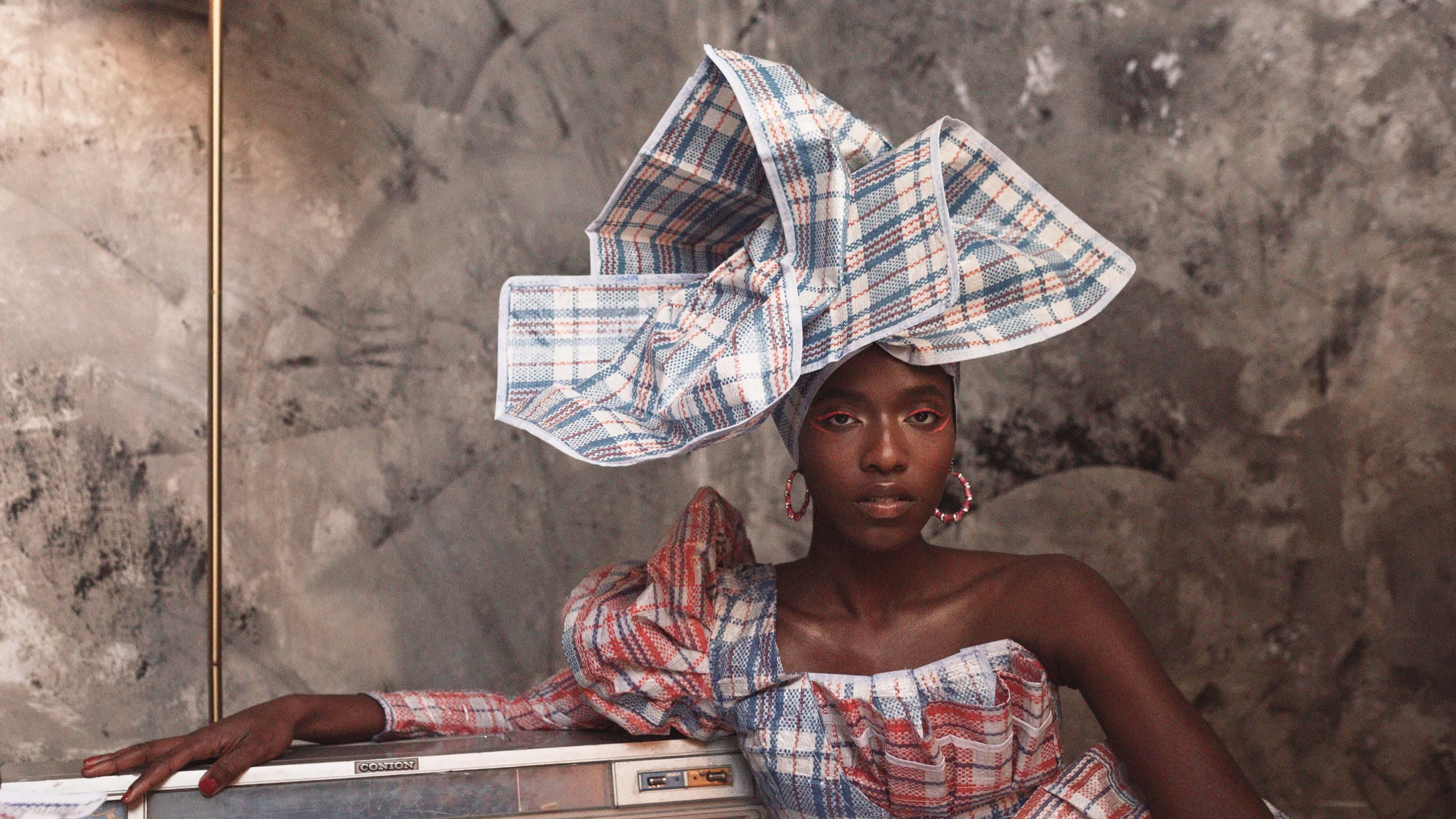 A woman wears a plaid head wrap and dress and poses near a transistor radio