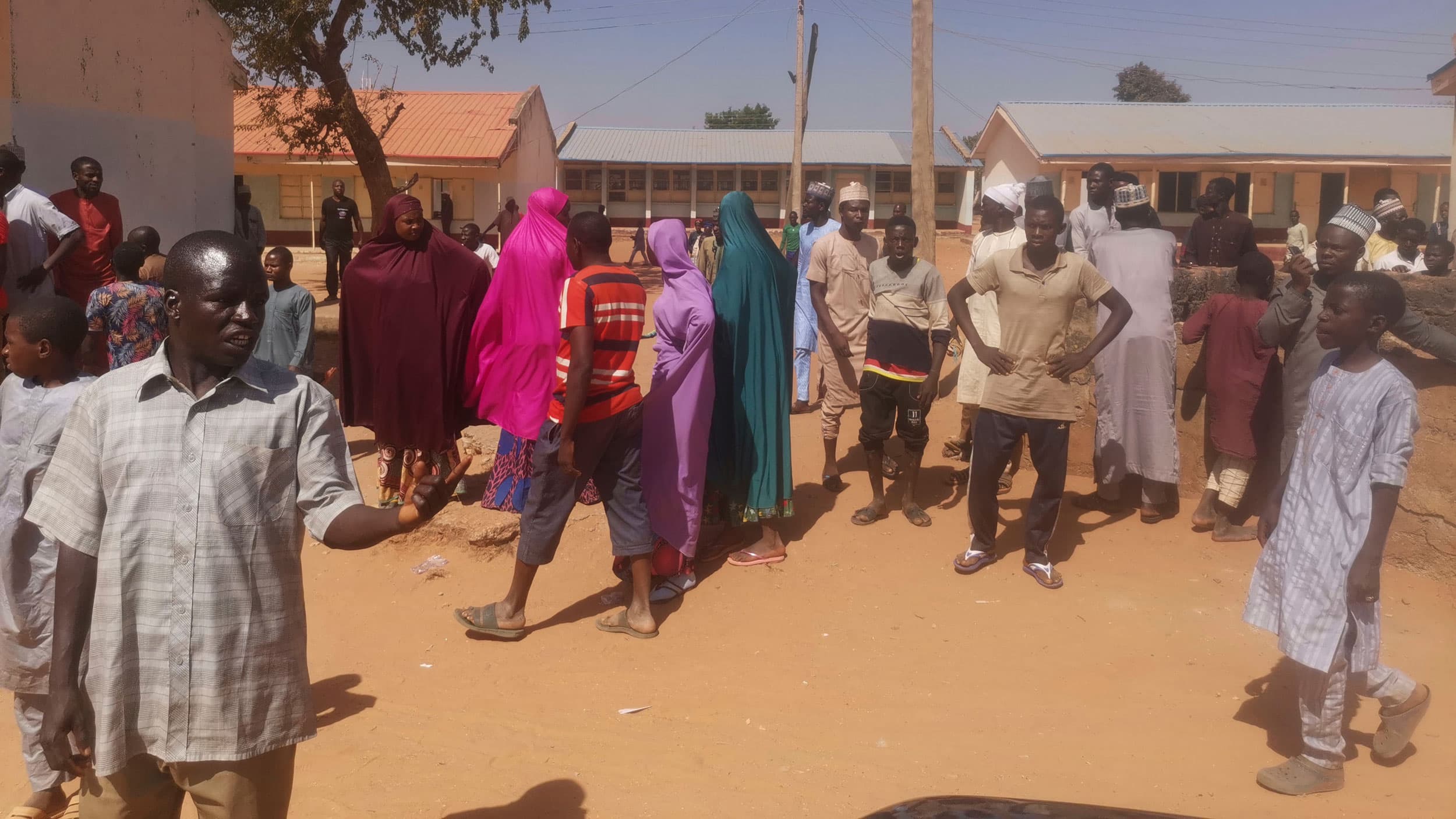 People are shown gathering in a dusty courtyard with several single story buildings in the background.