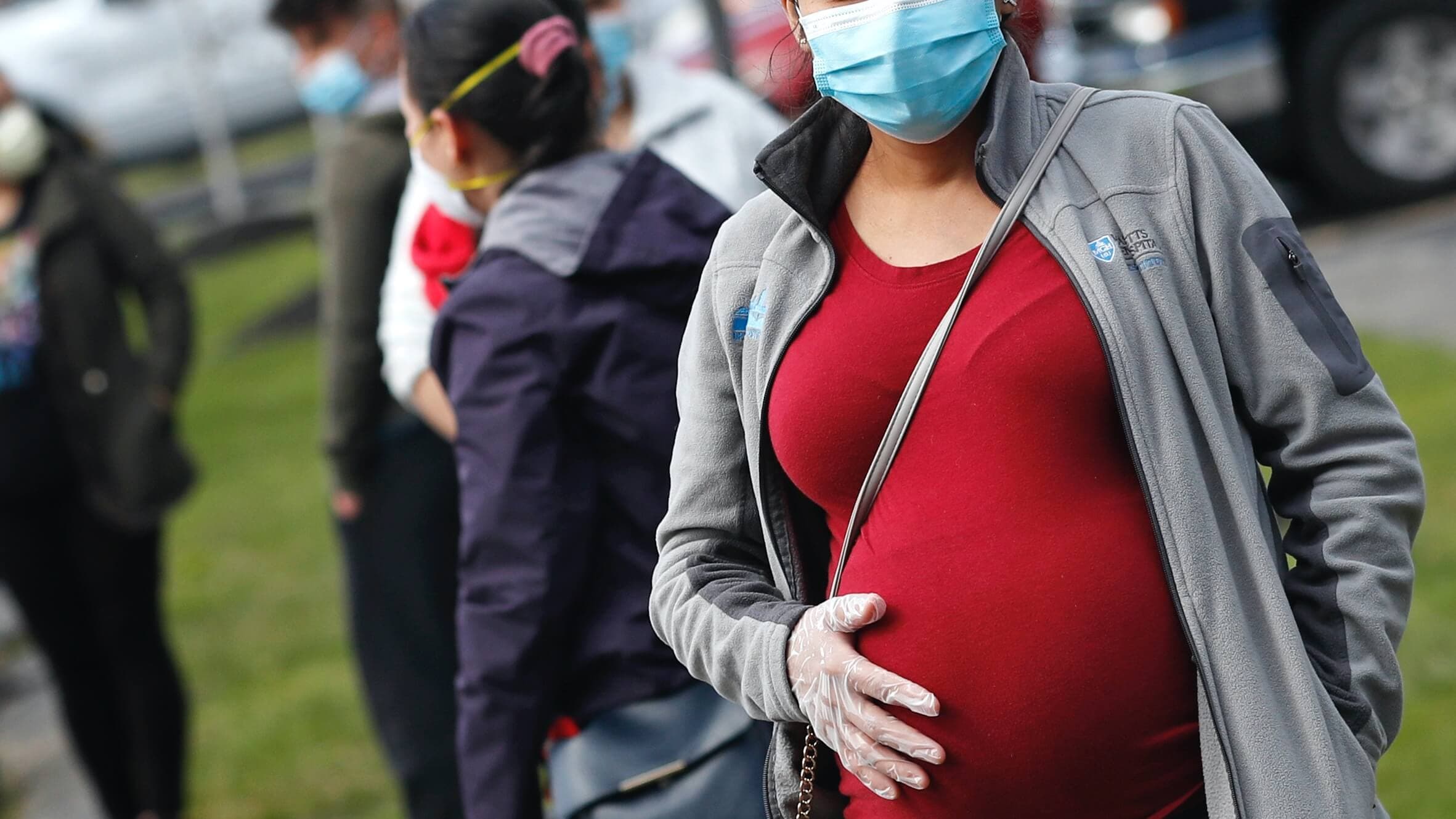 A pregnant woman wearing a red shirt holds her belly with a plastic gloved hand.