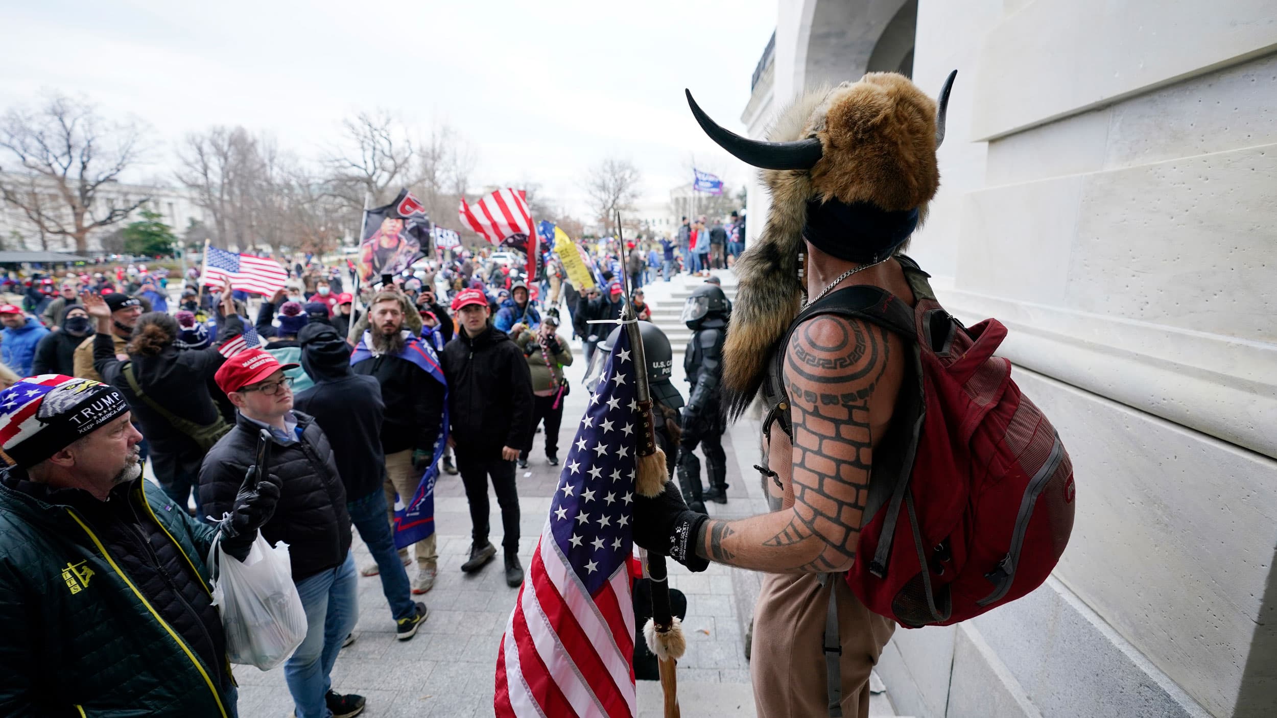 A man wearing a fir hat with horns and holding a US flag is shown standing among a large crowd of Trump supporters.