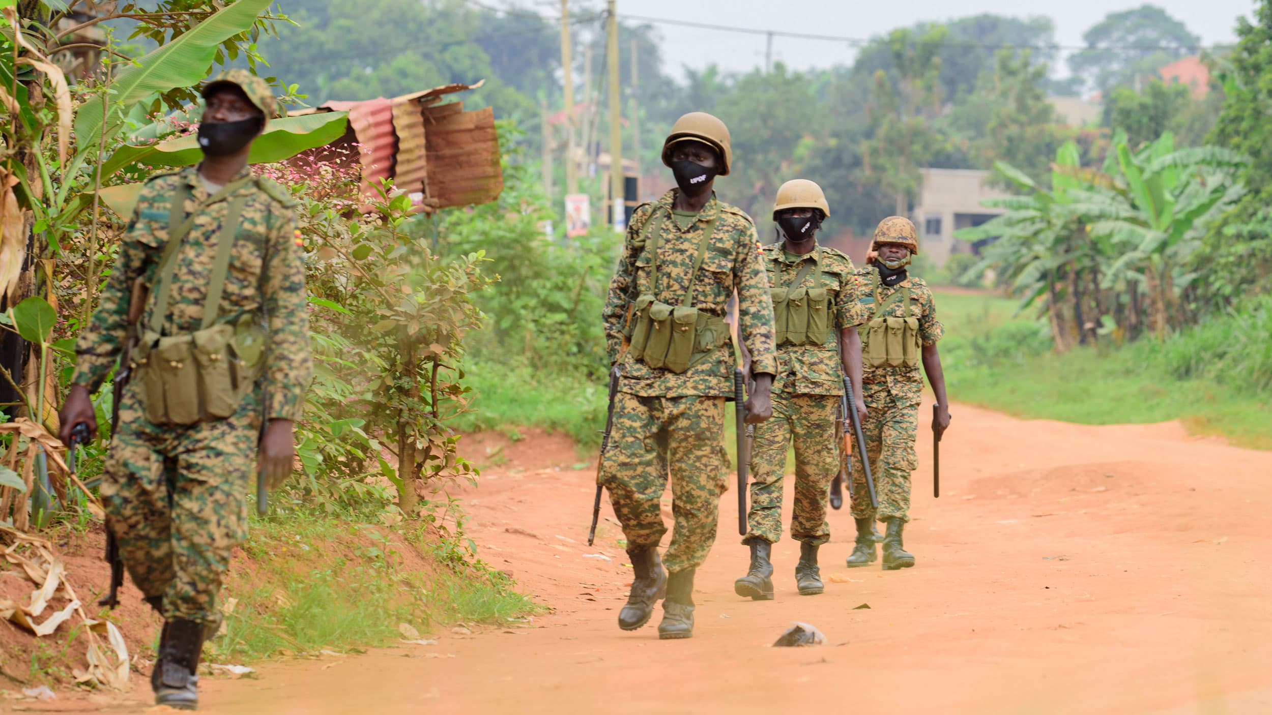 Four soldiers are shown wearing military fatigues and helmets while carrying weapons and walking in a dirt road.