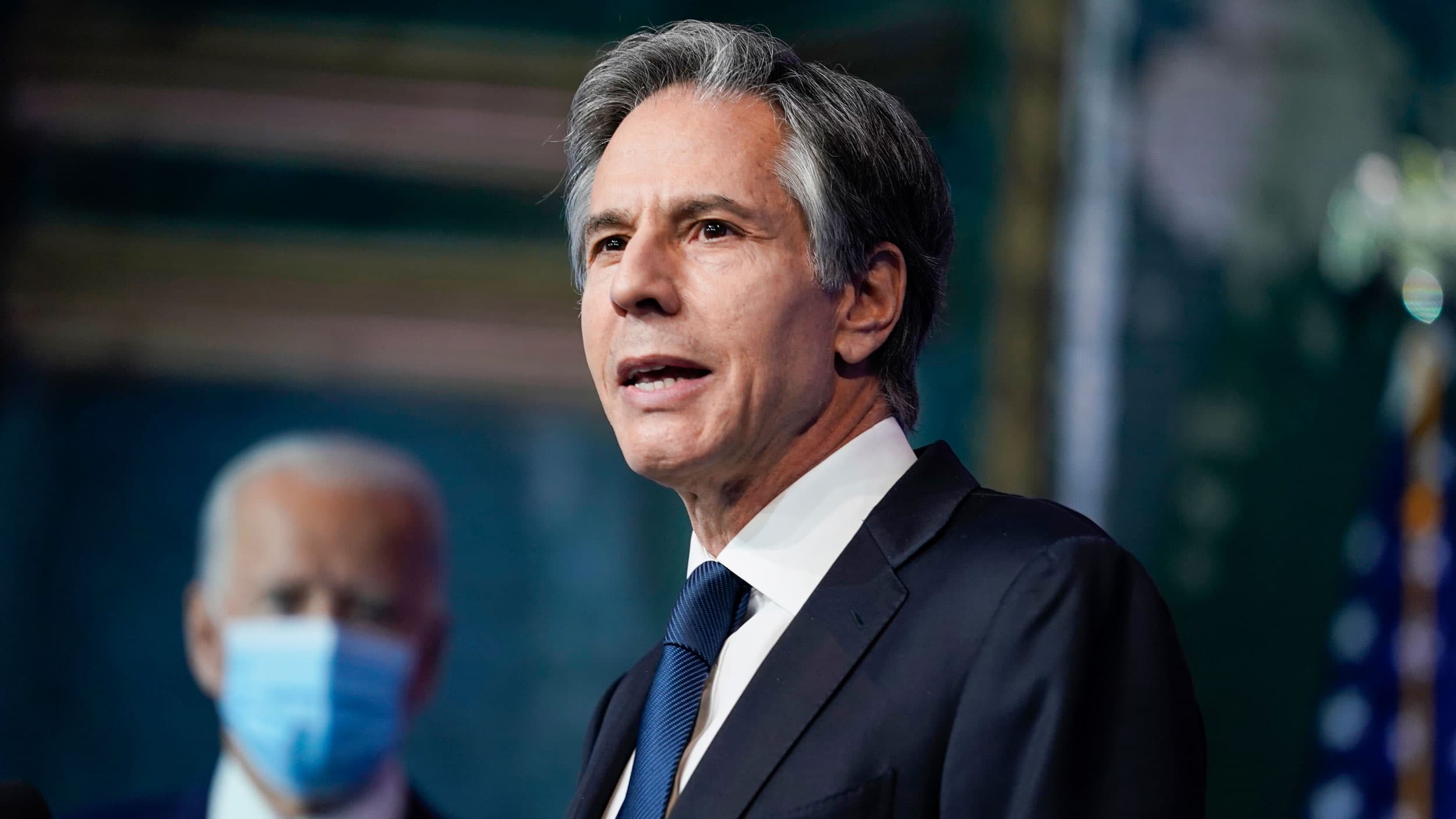 Tony Blinken is shown wearing a blue suit and tie while speaking with Joe Biden wearing a face mask in the distance.