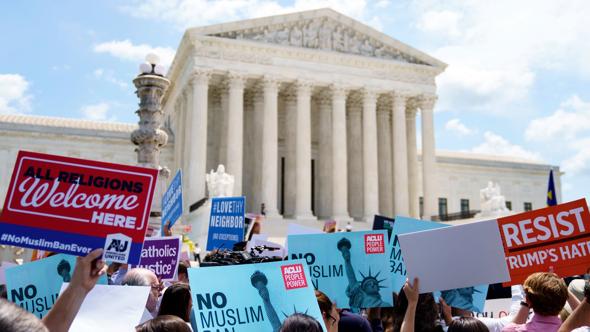 A group of people are shown holding different placards protesting the Trump administration's travel ban with the US Supreme Court building in the distance.