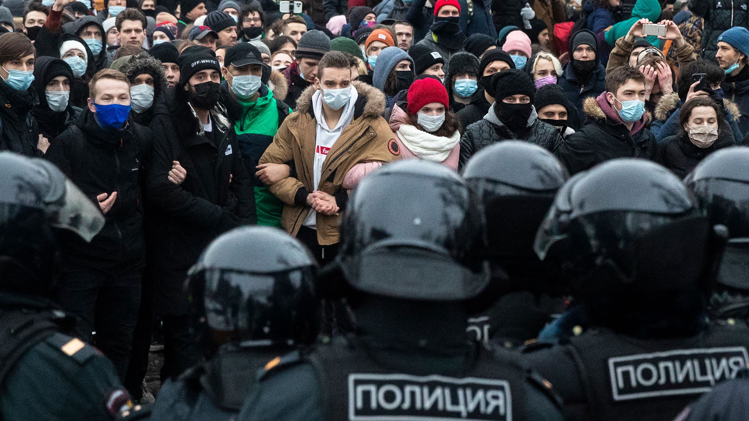 A large crowd of people are shown with arms locked standing opposite a line of police wearing riot gear with black helmets.