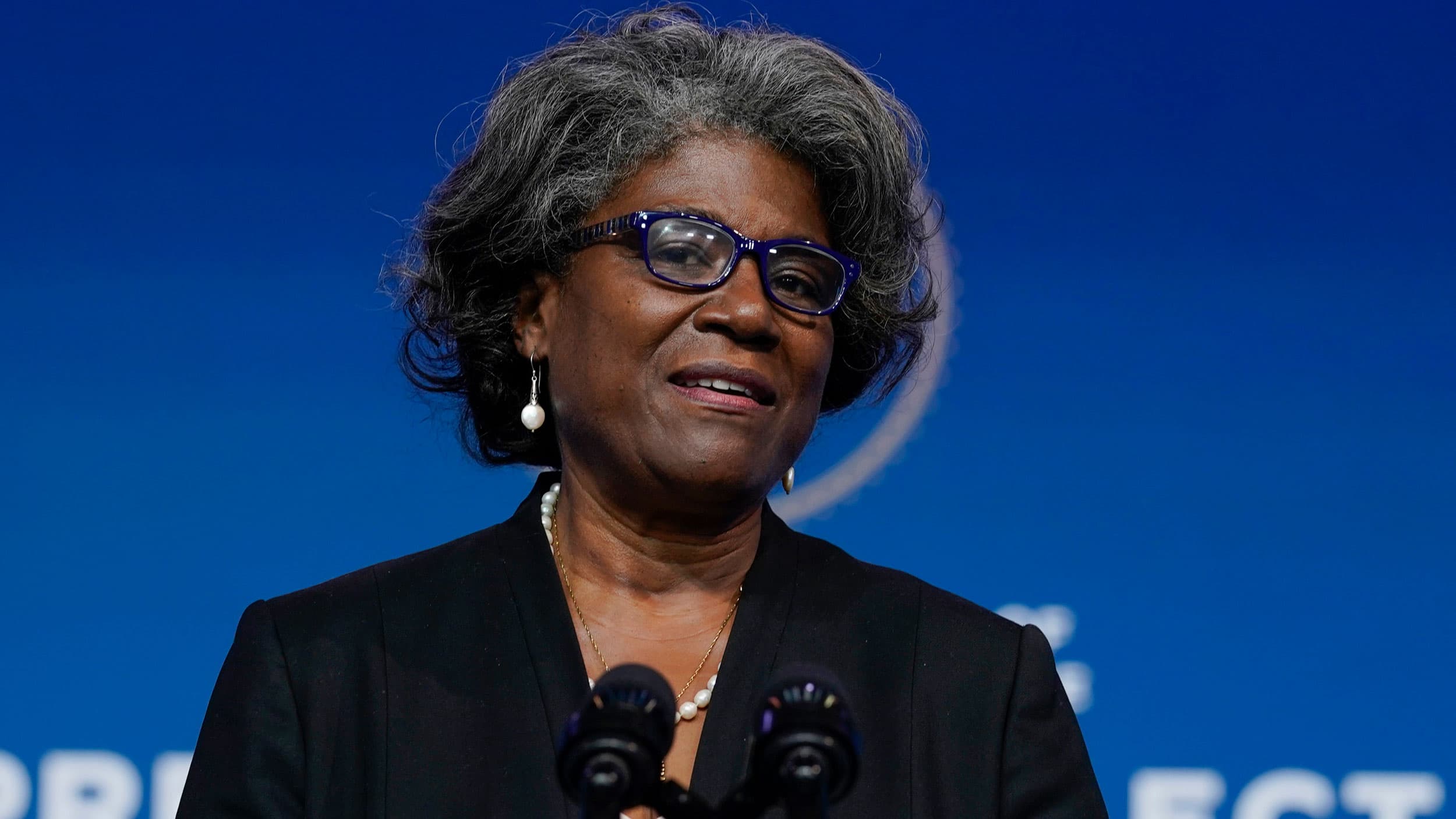 Linda Thomas-Greenfield is shown speaking at a podium with microphones in front of her and a blue backgroud in the distance.