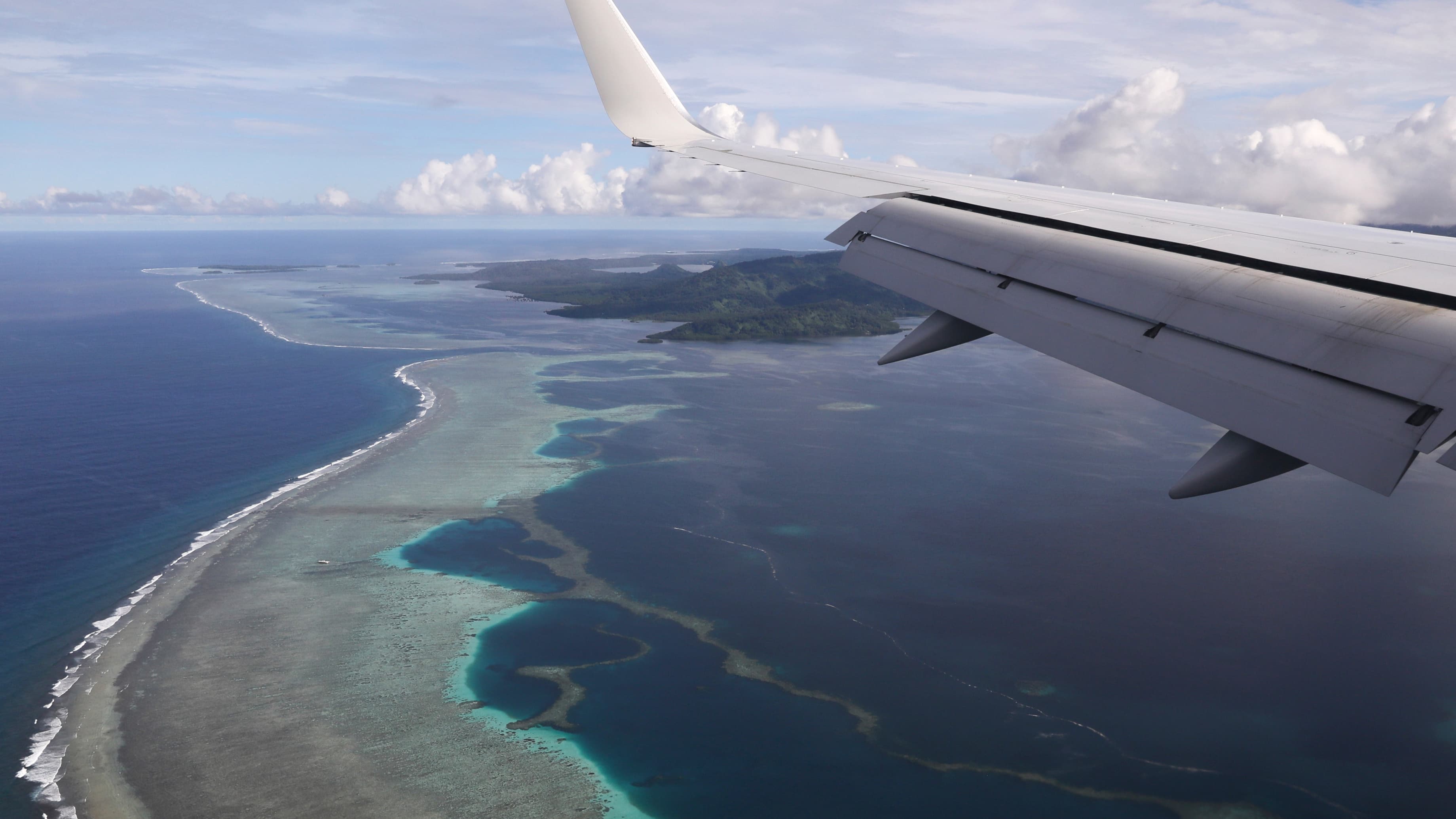 A plane flies over blue ocean waters and small island strips.