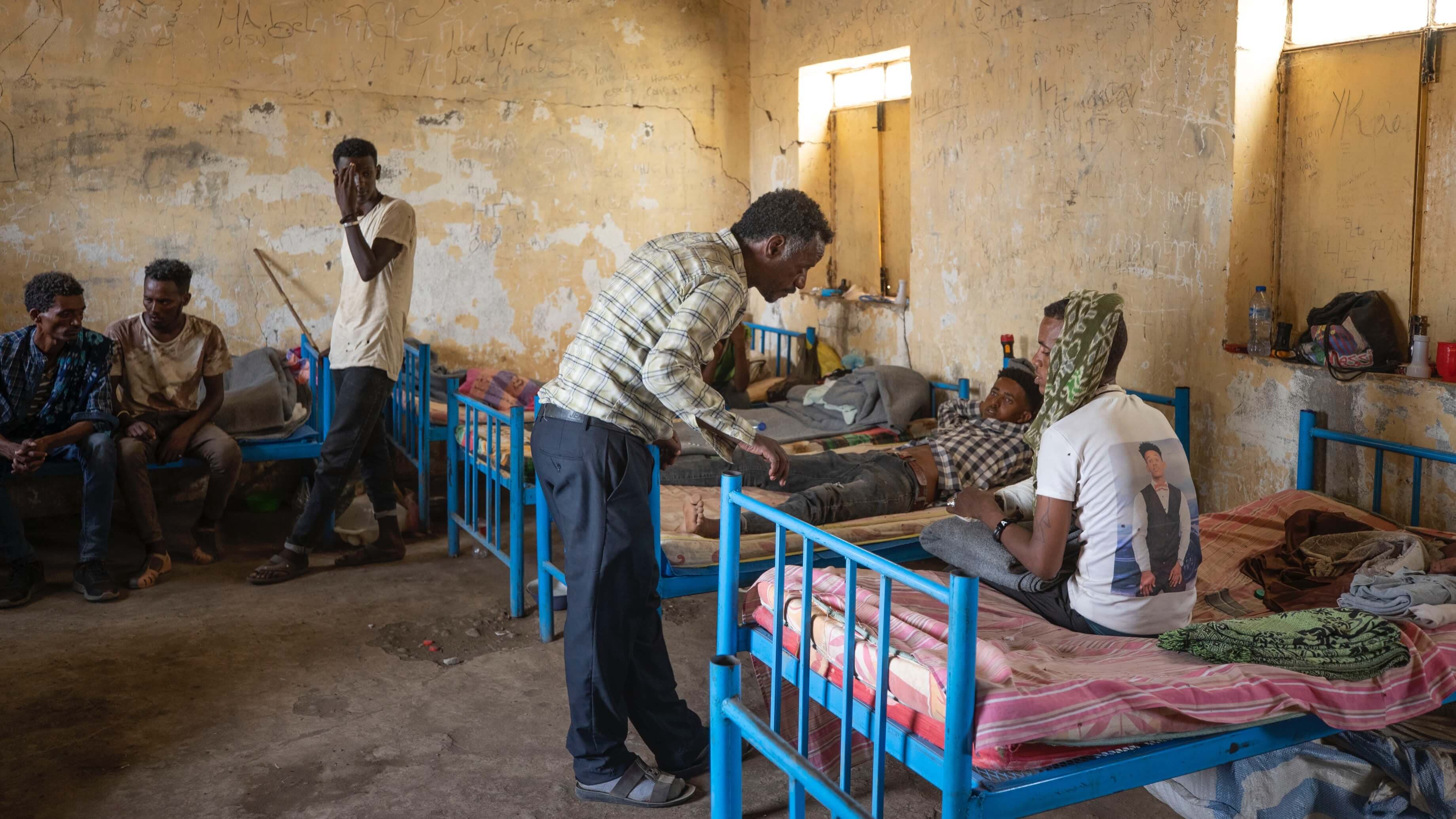 A doctor wearing a plaid shirt and slacks leans over a patient in a blue metal bed.