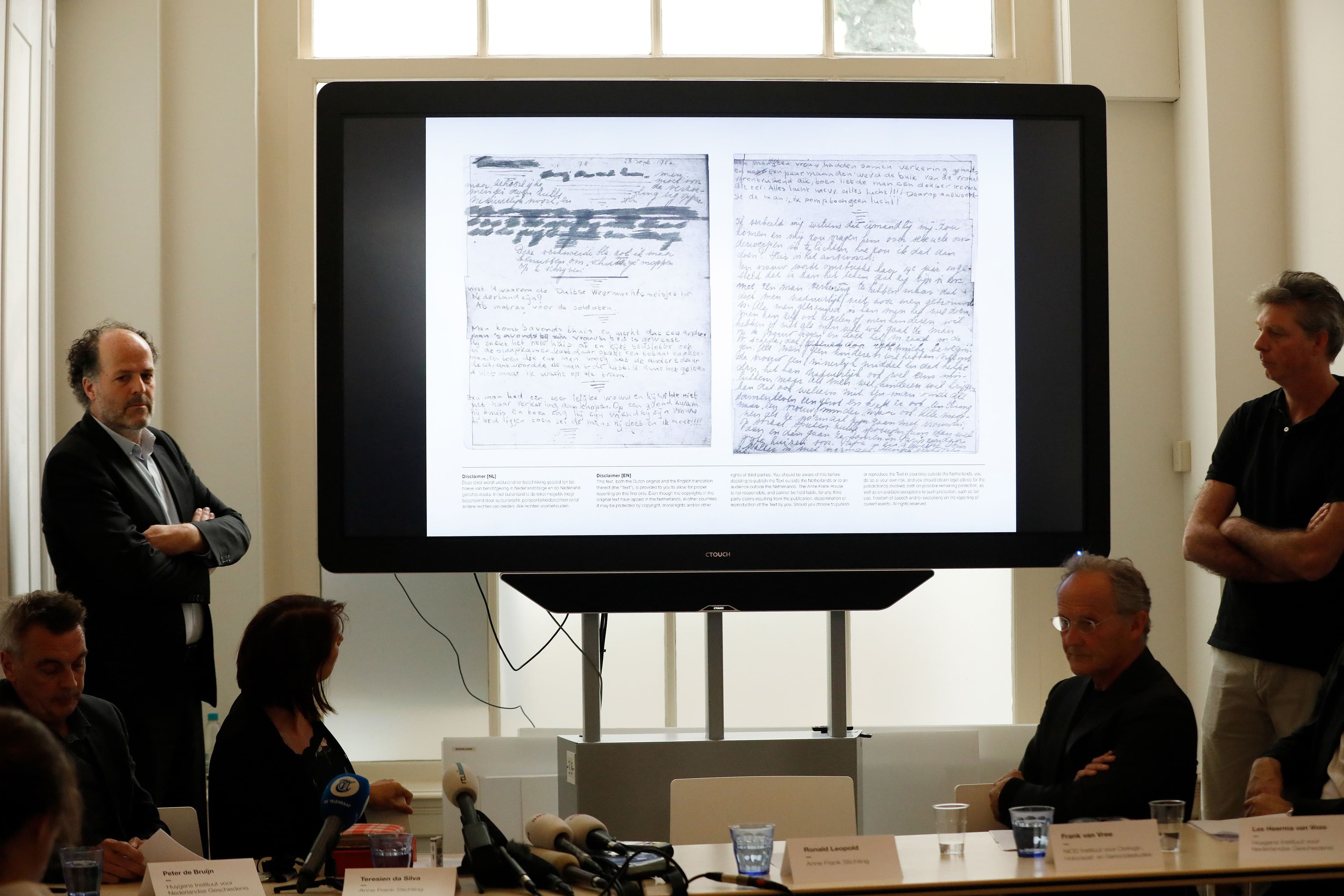 Two men stand on both sides of a projector screen, displaying pages of Anne Frank's handwriting.