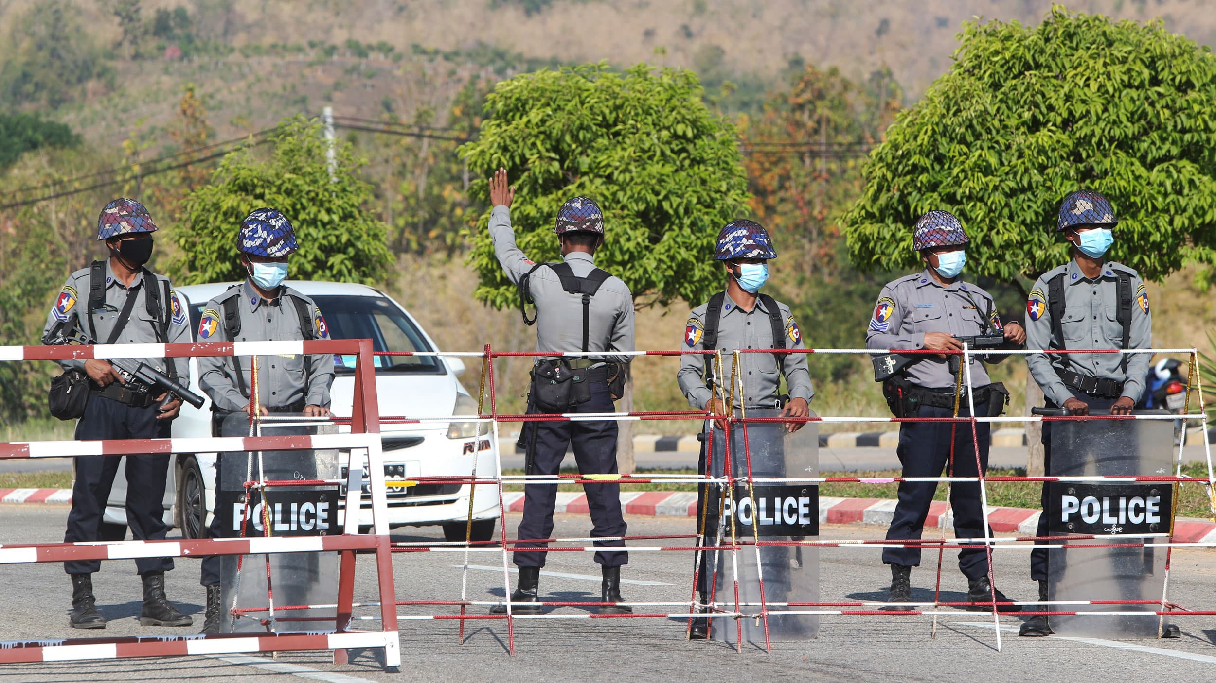 A line of six security authorities are shown standing next to a metal baracade blocking a road with one man holding his hand up stopping an approaching car.
