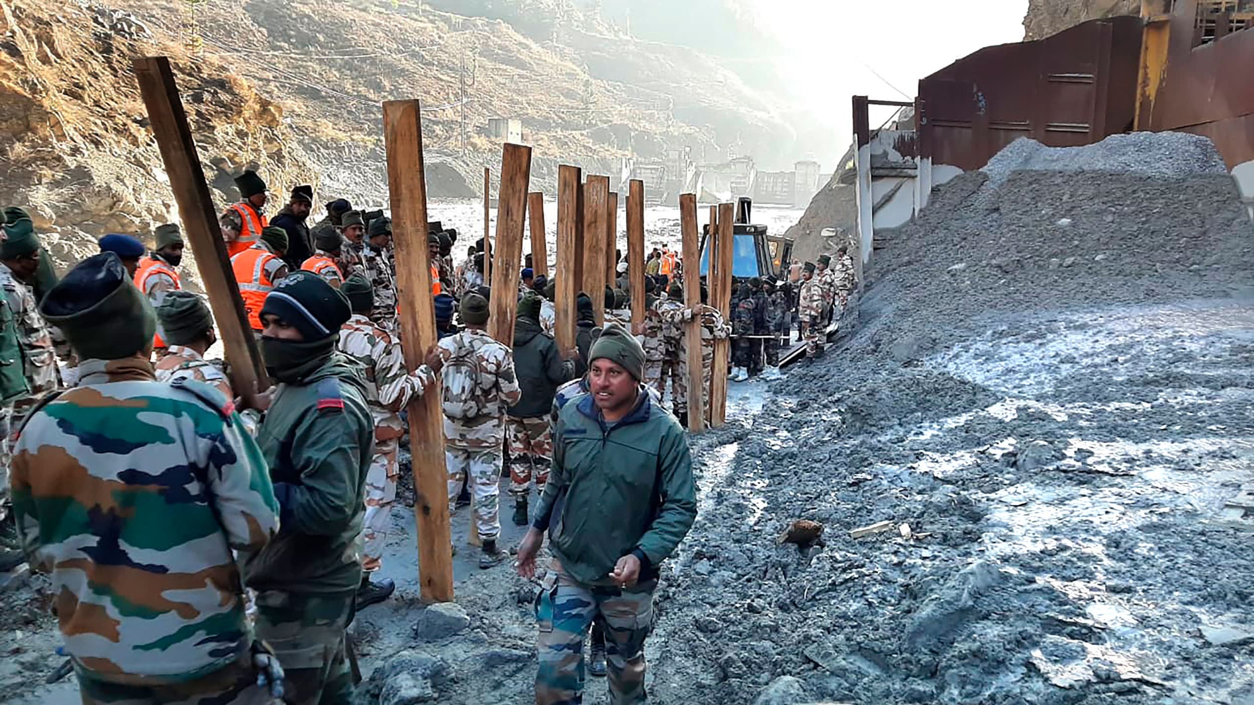 Several rescue workers are shown standing in the muddy collapse of a dam with several pieces of large wood sticking up from the ground.