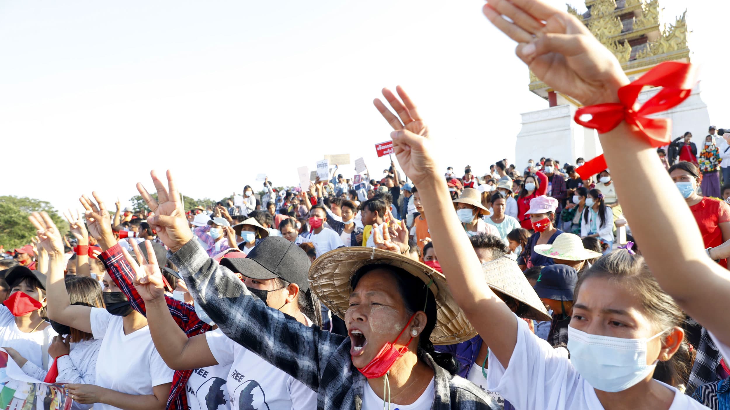 A large crowd of people are shown, many with their hands raise and holding three fingers in the air with a golden-color shrine seen in the distance.