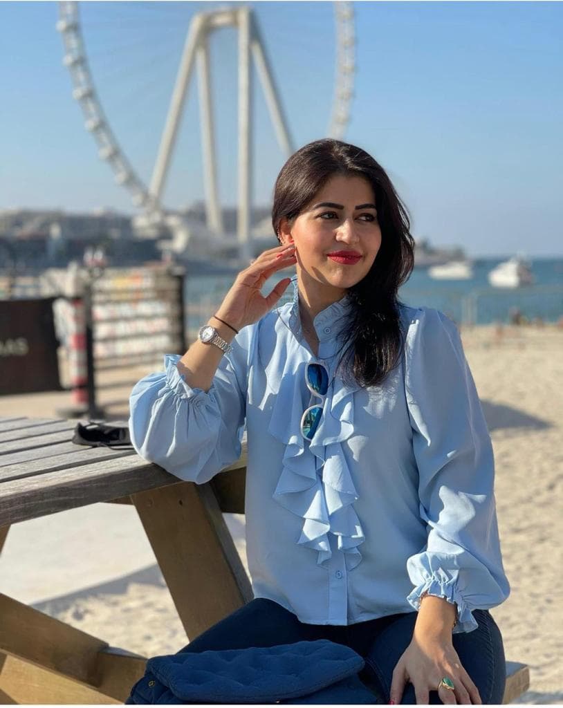 A woman wearing a blue top and jeans poses on a bench outdoors in front of a Ferris wheel.