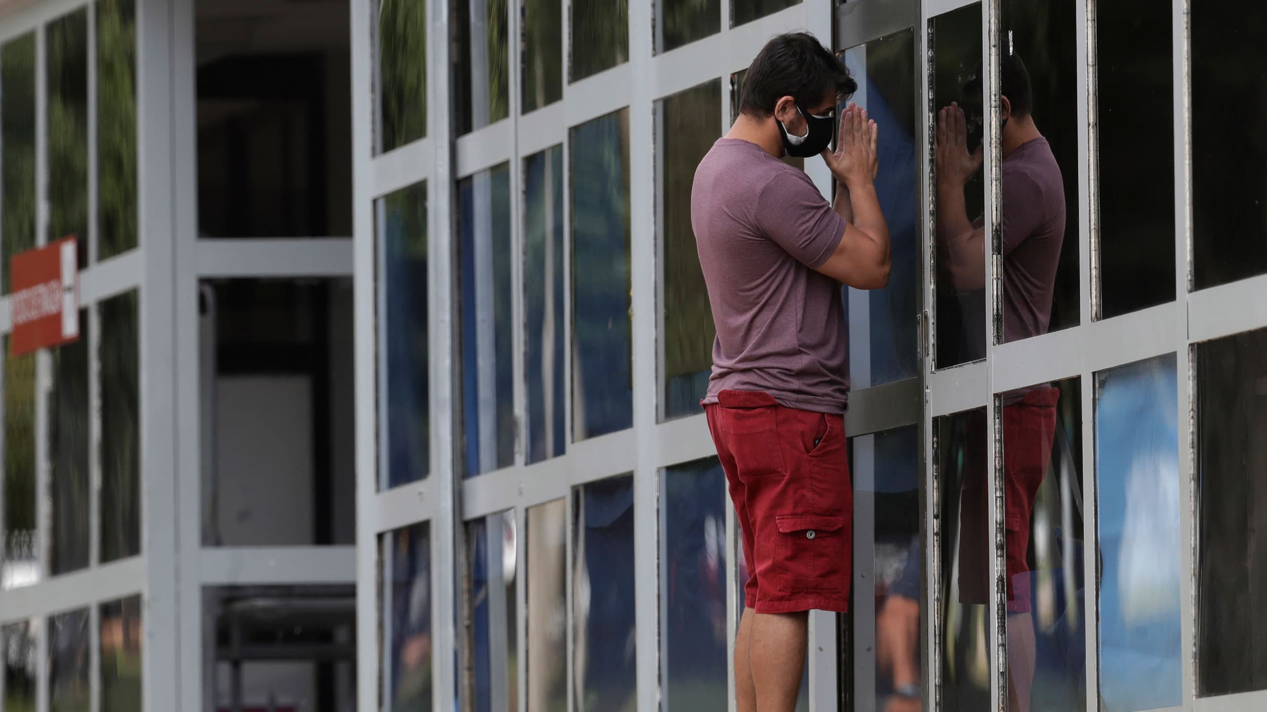 A man is shown wearing shorts and a t-shirt praying outside the windows of an improvised ICU.