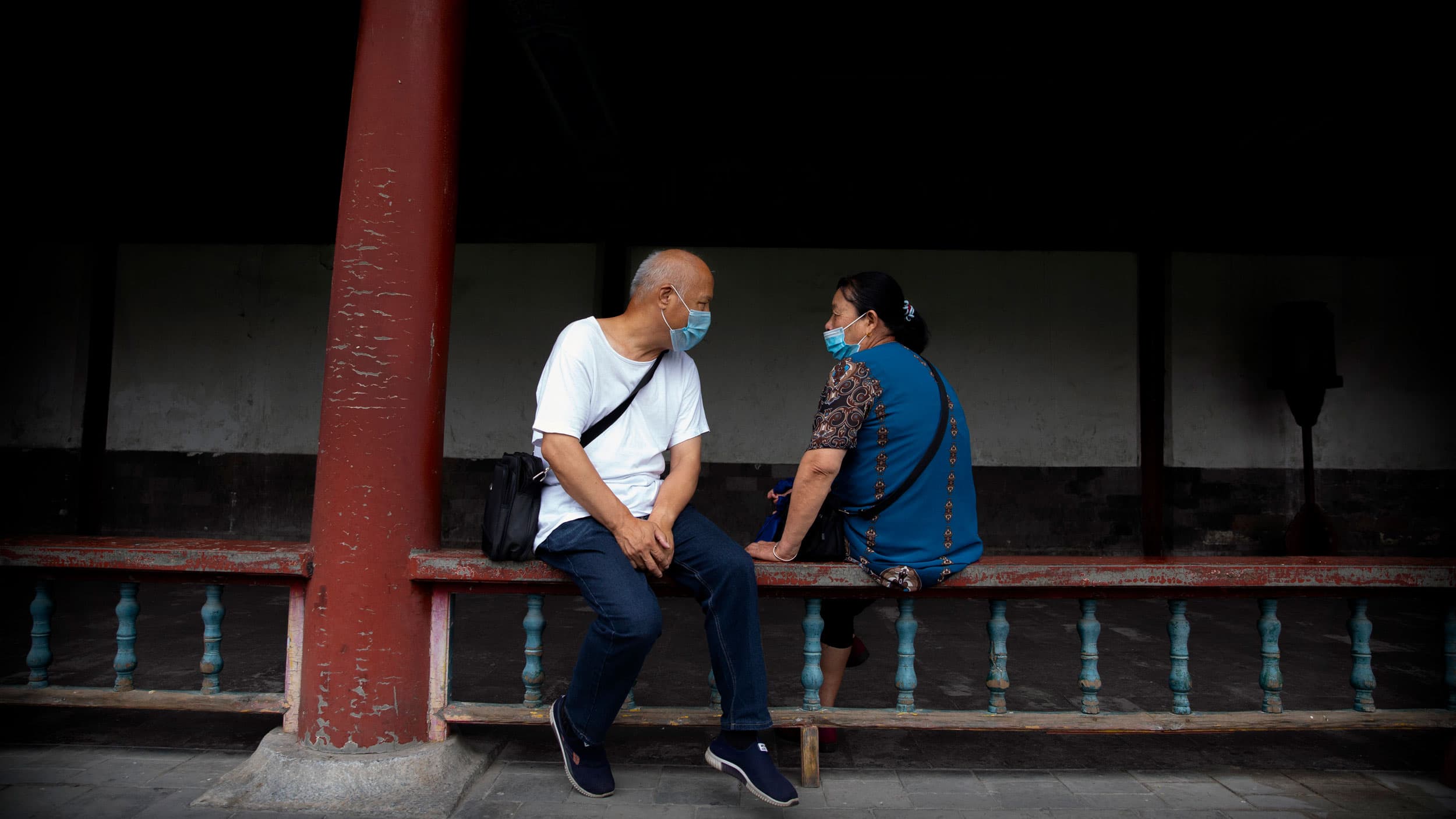 A man and woman are shown sitting on either side of a wooden railing and facing each other while wearing protective masks.