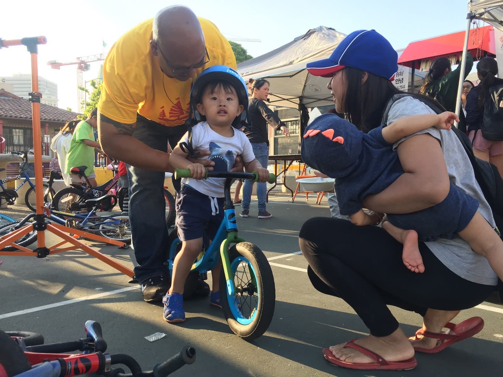 A man wearing a yellow shirt helps a child on a bike with his family members kneeling down near him.