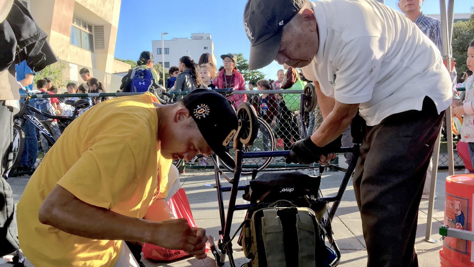 A man in a yellow shirt helps fix the bike of an elder leaning over bike wearing casual clothes and cap