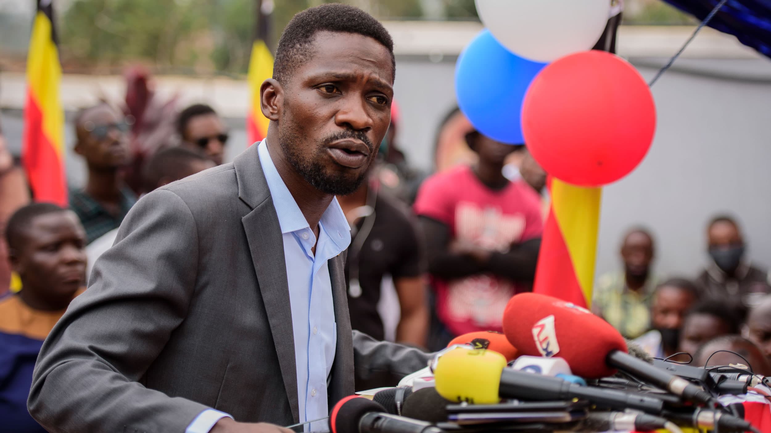 Ugandan opposition figure Bobi Wine is shown wearing a gray blazer an speaking into severla microphones.