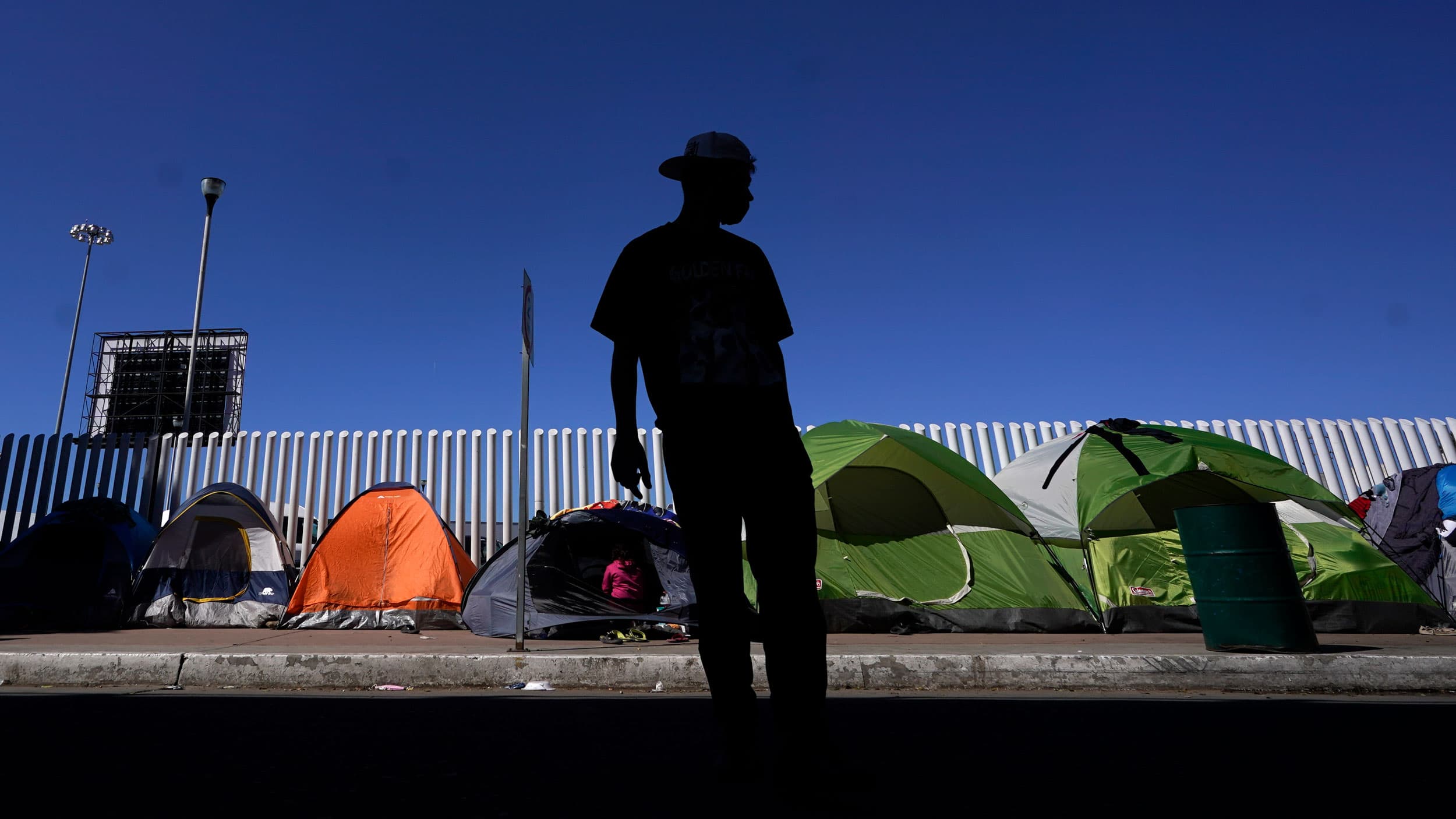 A man is shown in shadow and wearing a brimmed hat with several pup tents shown in focus in the background.