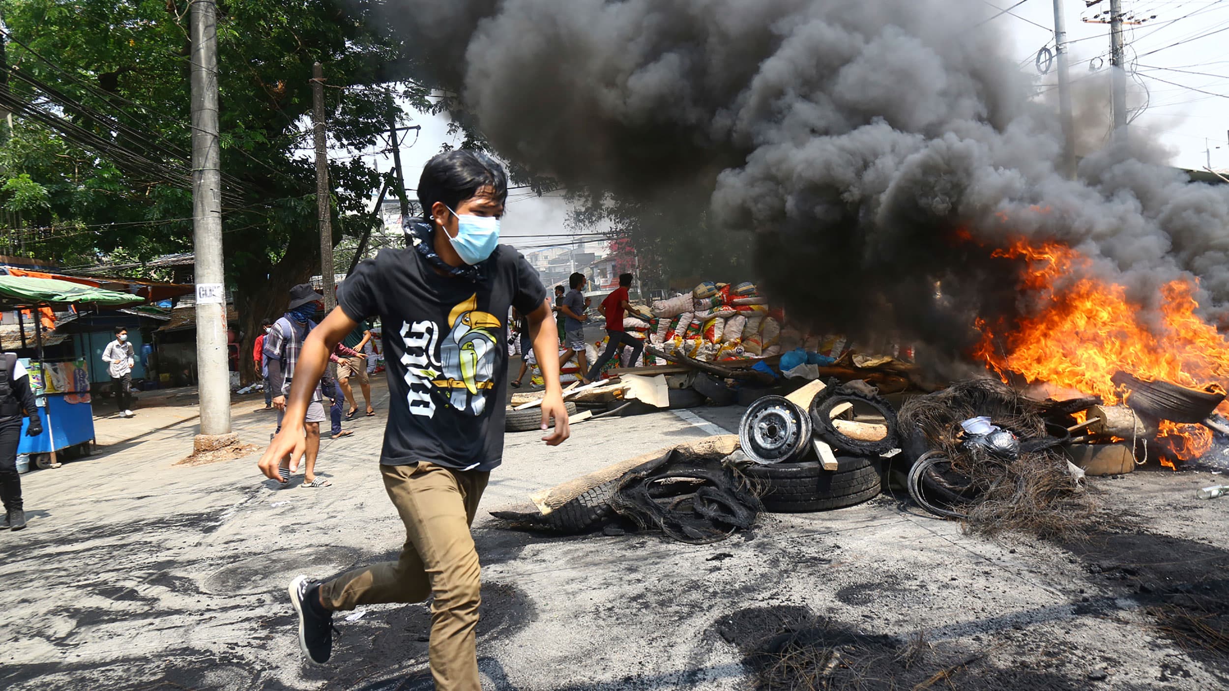 A man is shown wearing a medical mask while running past a large fire in the street behind him.