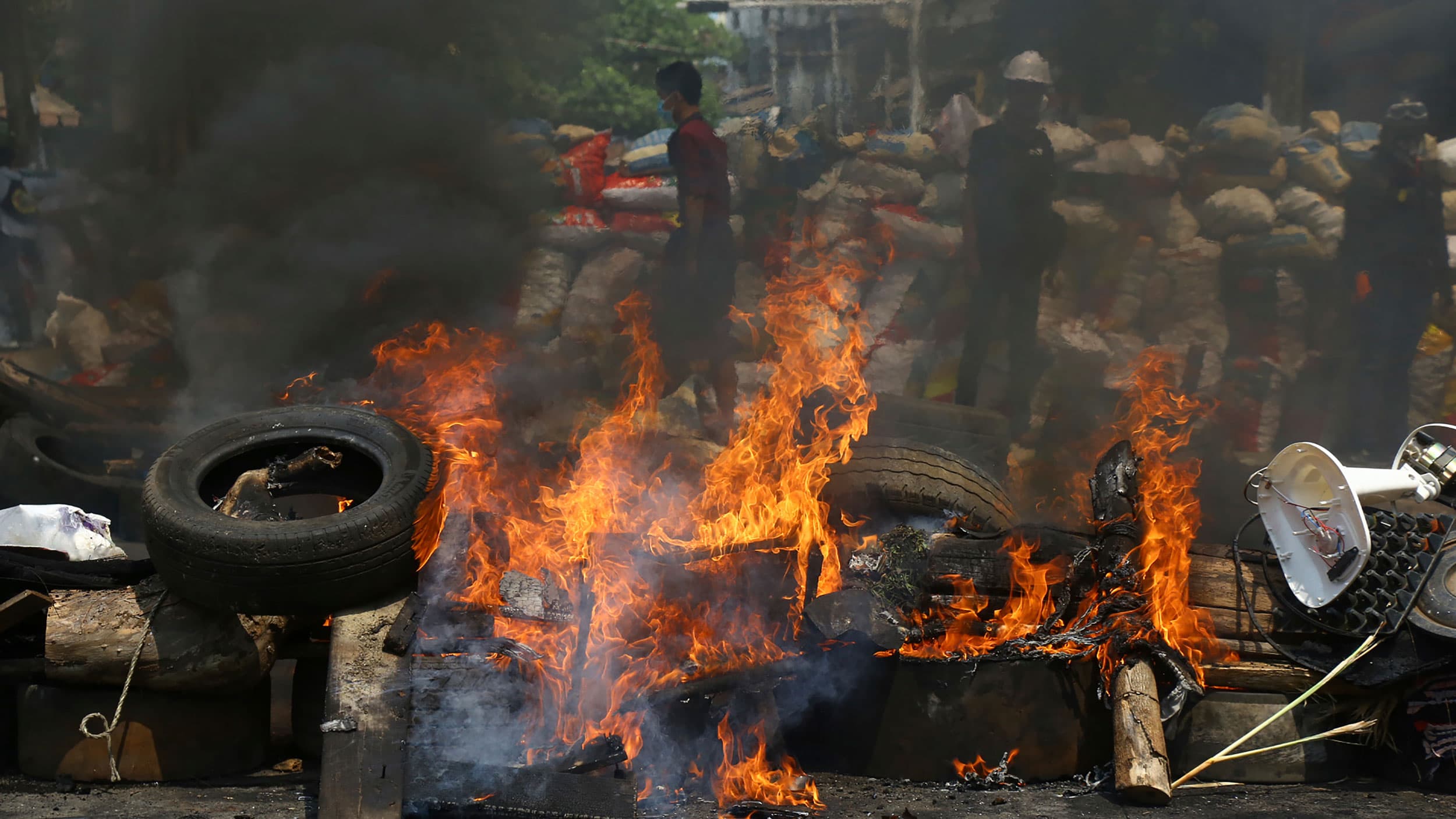 A large fire is shown with dark smoke rising and protesters shown in the distance.