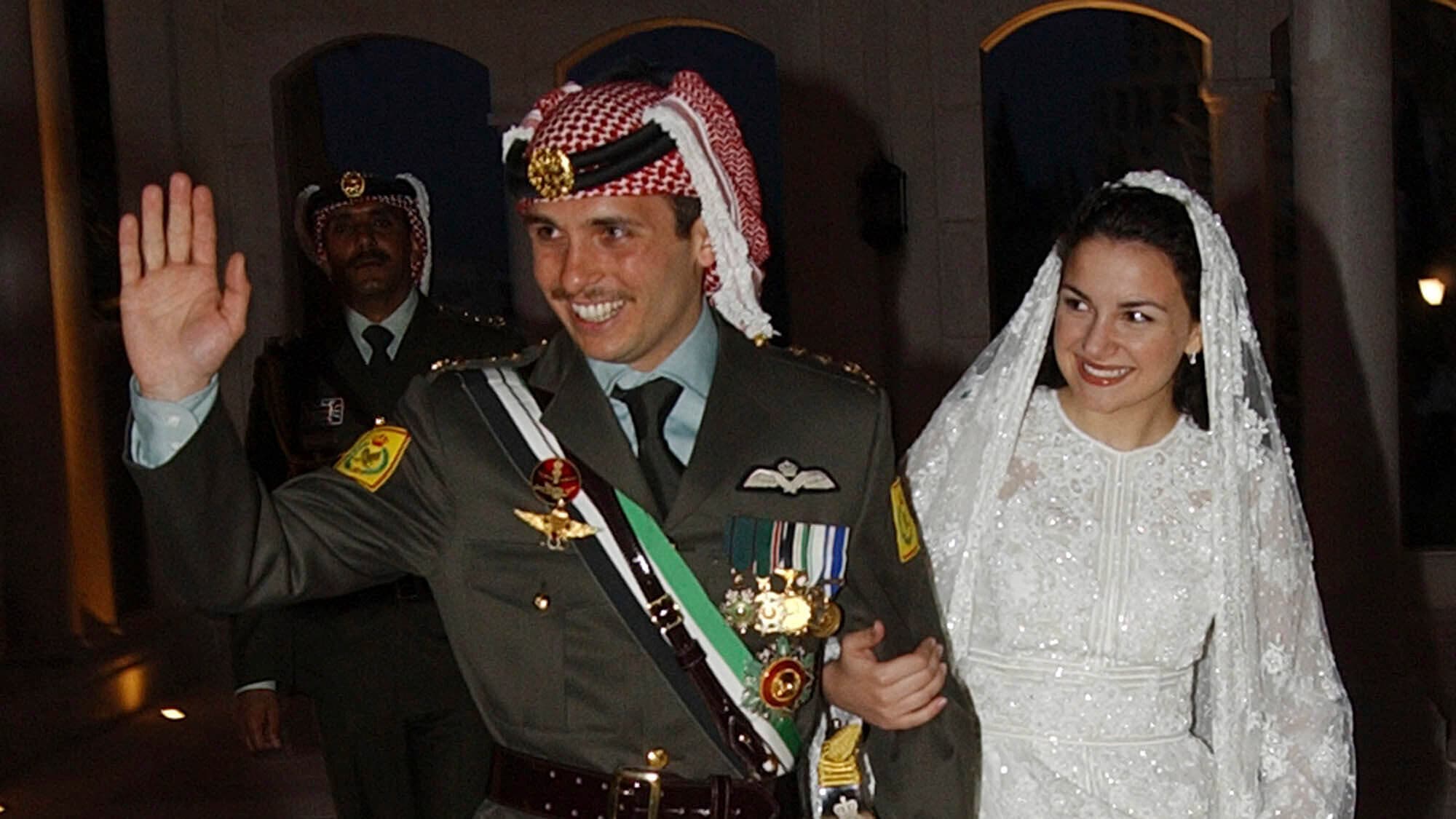Prince Hamzah waves, wearing a military uniform walks with his bride, Prince Noor, wearing a white wedding dress and they both smile.