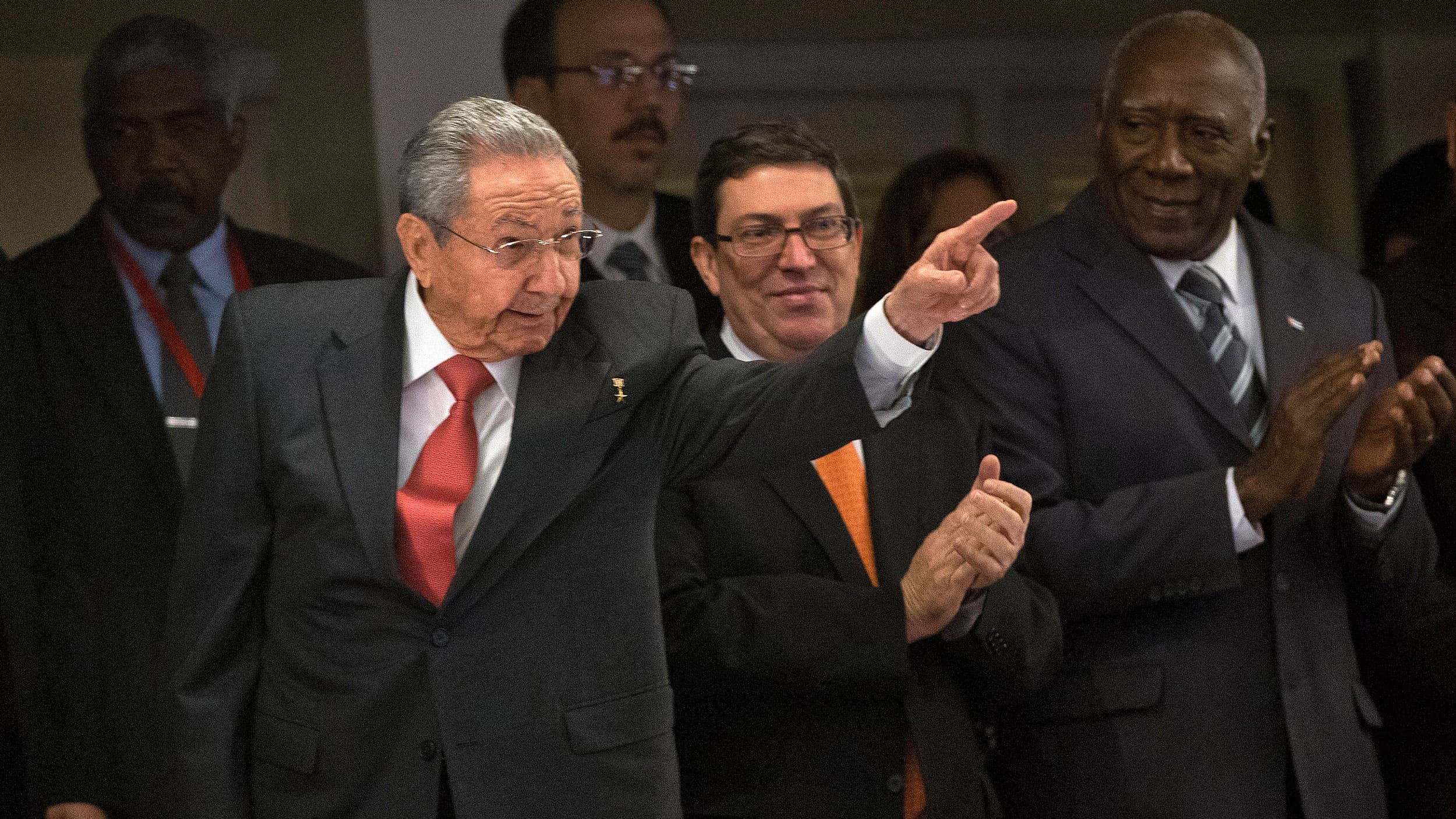 Raúl Castro is shown wearing a dark suit and red tie with his left arm raised and finger pointing.
