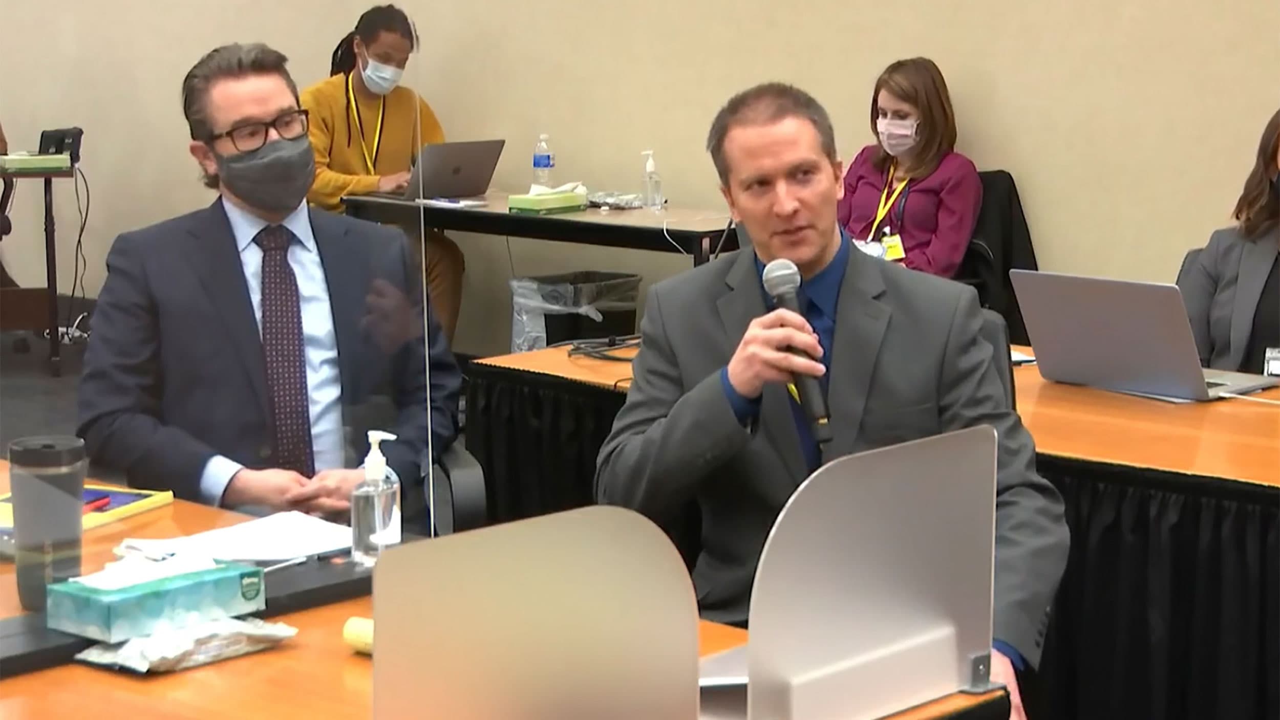 Derek Chauvin in gray suit hold microphone as he speaks, while sitting next to his lawyer
