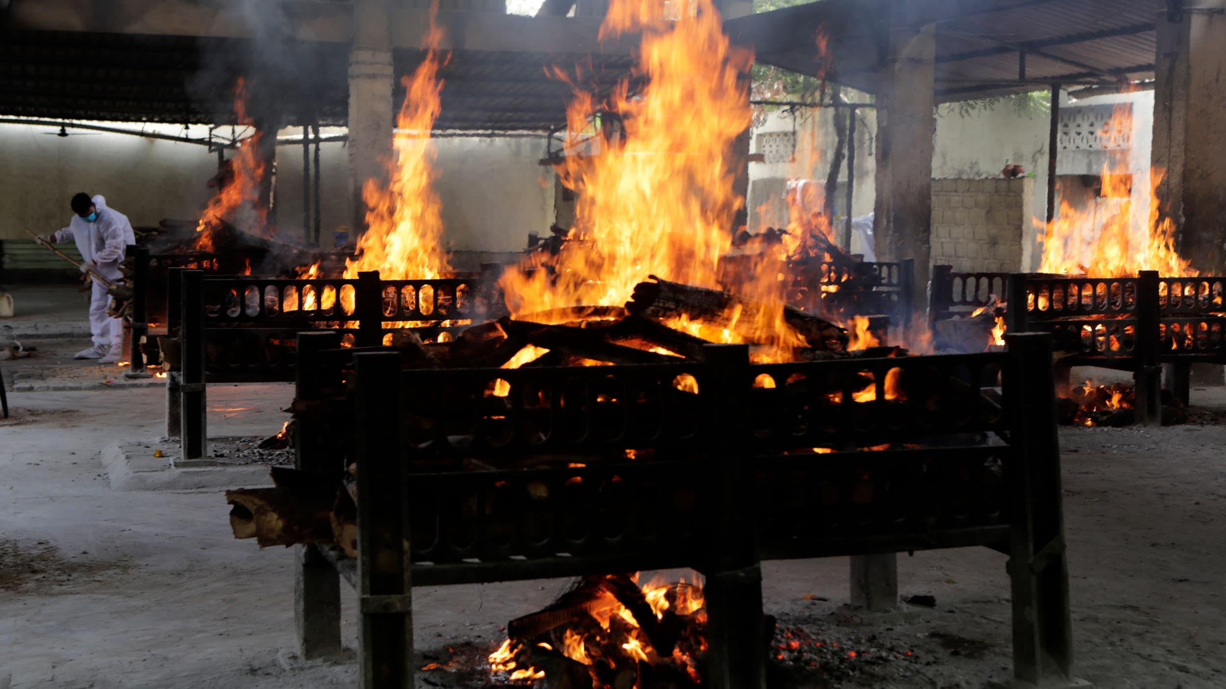 A row of cremation pyres are shown with large orange flames and a worker wearing white off in the distance.