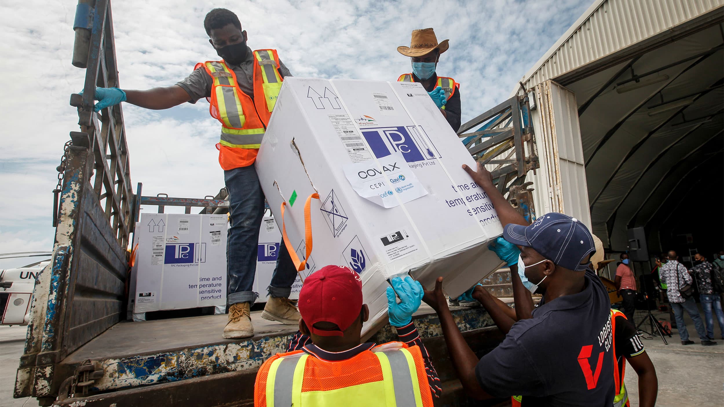 Men in orange and yellow jackets lift a box of vaccines from COVAX off of a truck bed