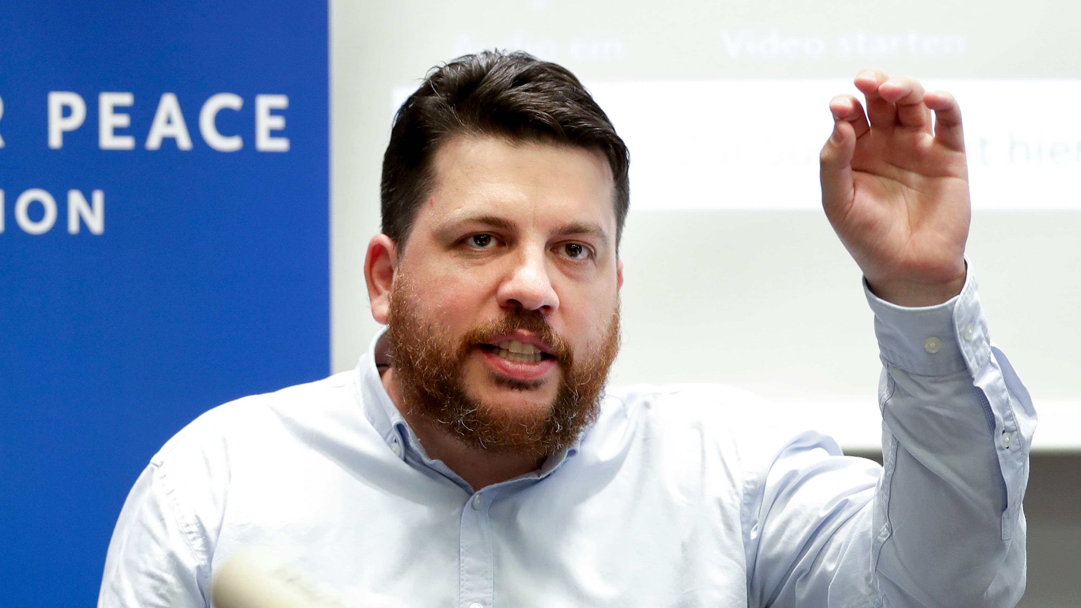 Leonid Volkov wears a white shirt and raises his hand in the air while speaking in front of a blue and white background.