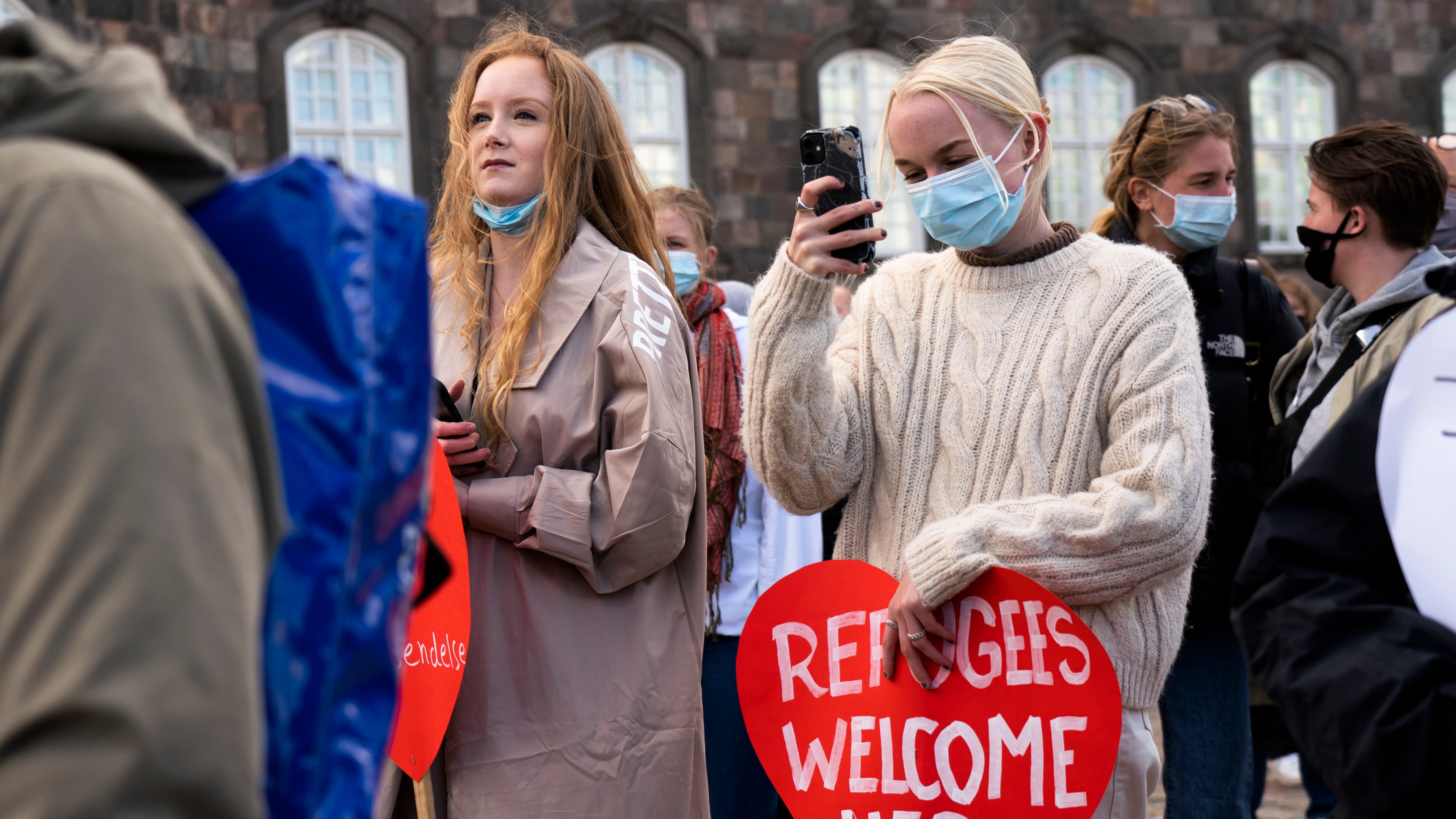 A group of people protest and carry a heart-shaped sign that says "Refugees welcome here."