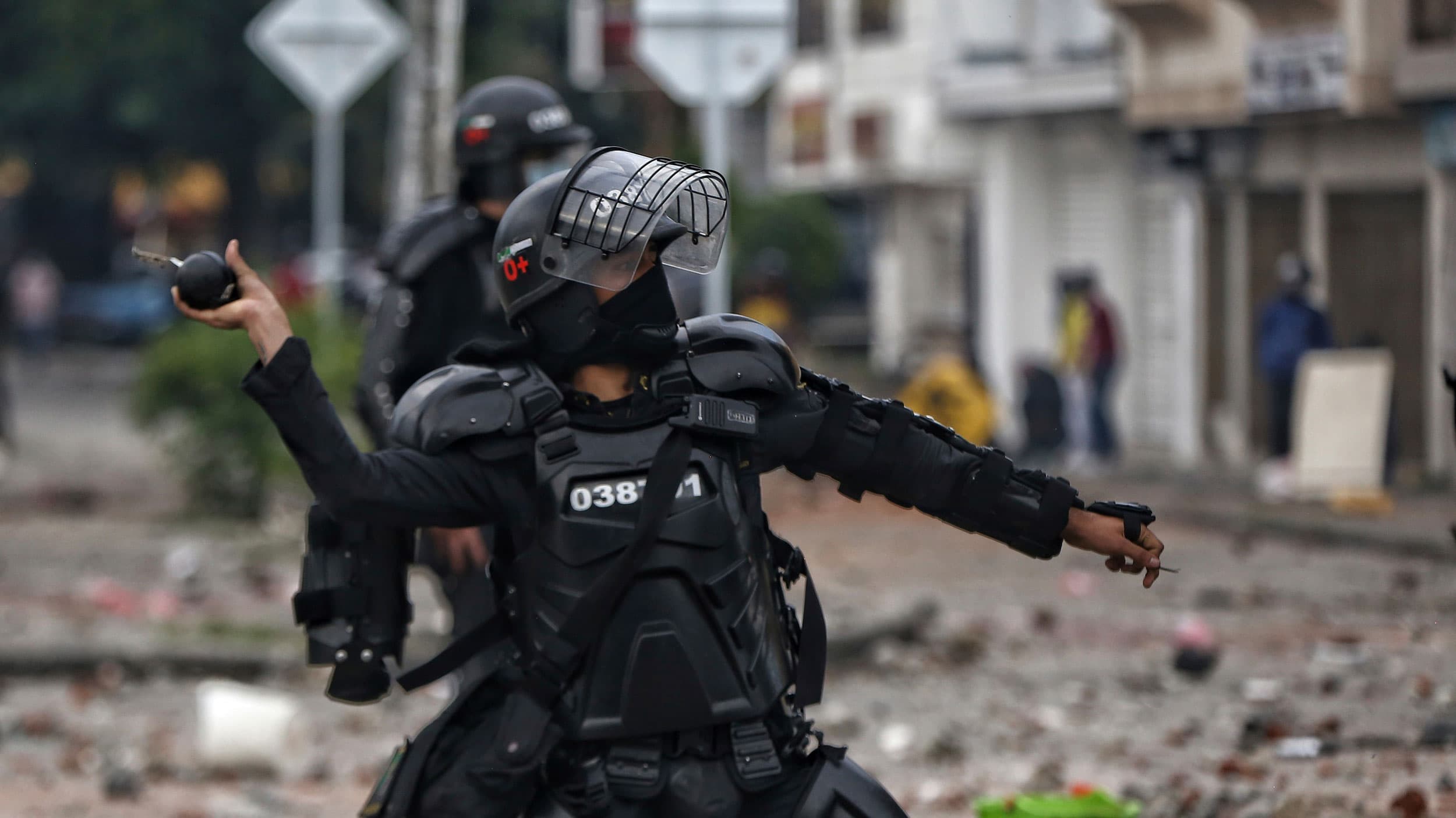 A police officer is shown wearing all black riot gear and face shield while throwing a stun grenade.