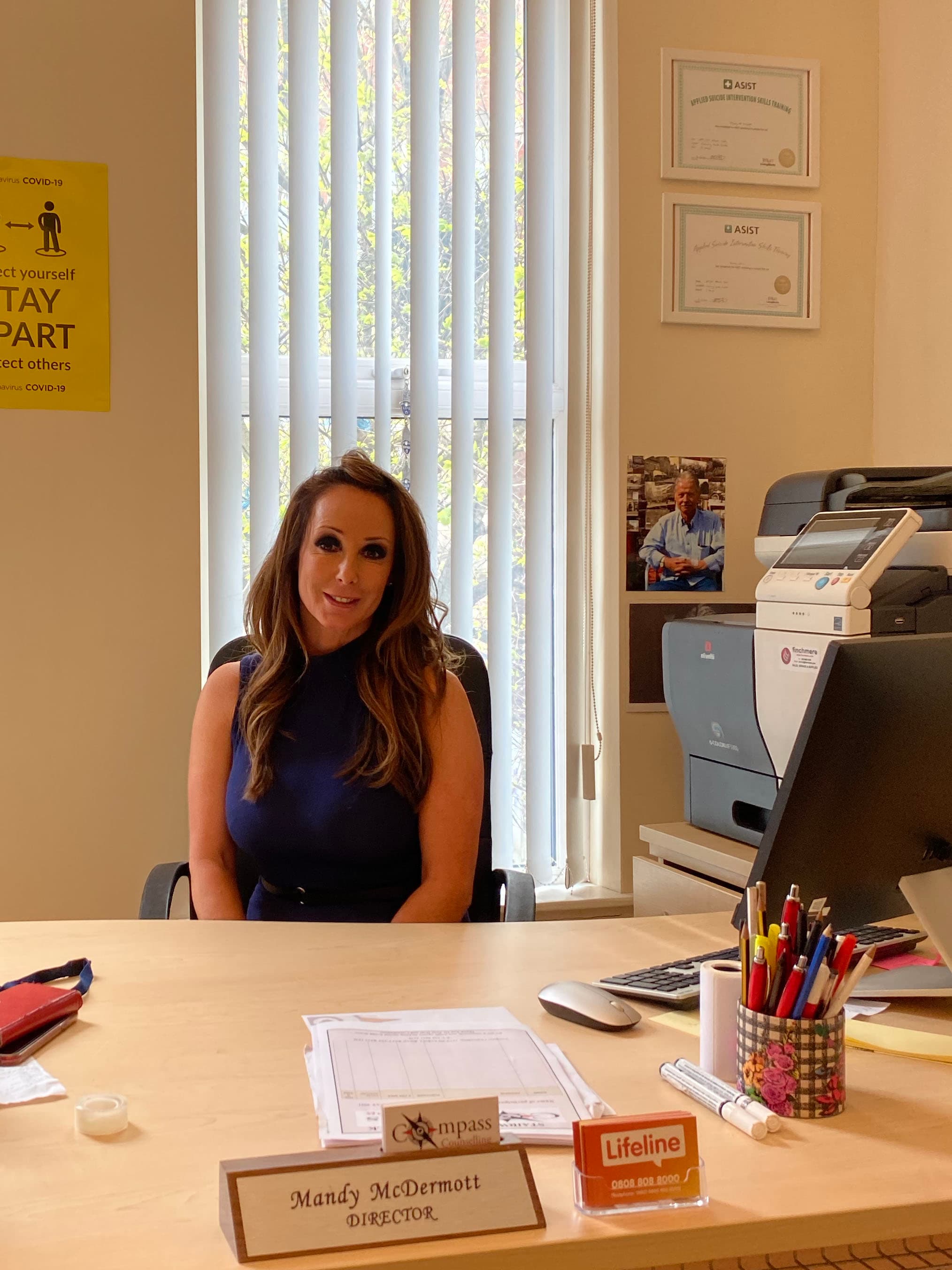 A white woman senior psychotherapist wears a black shirt and sits at a beige desk inside at her office.
