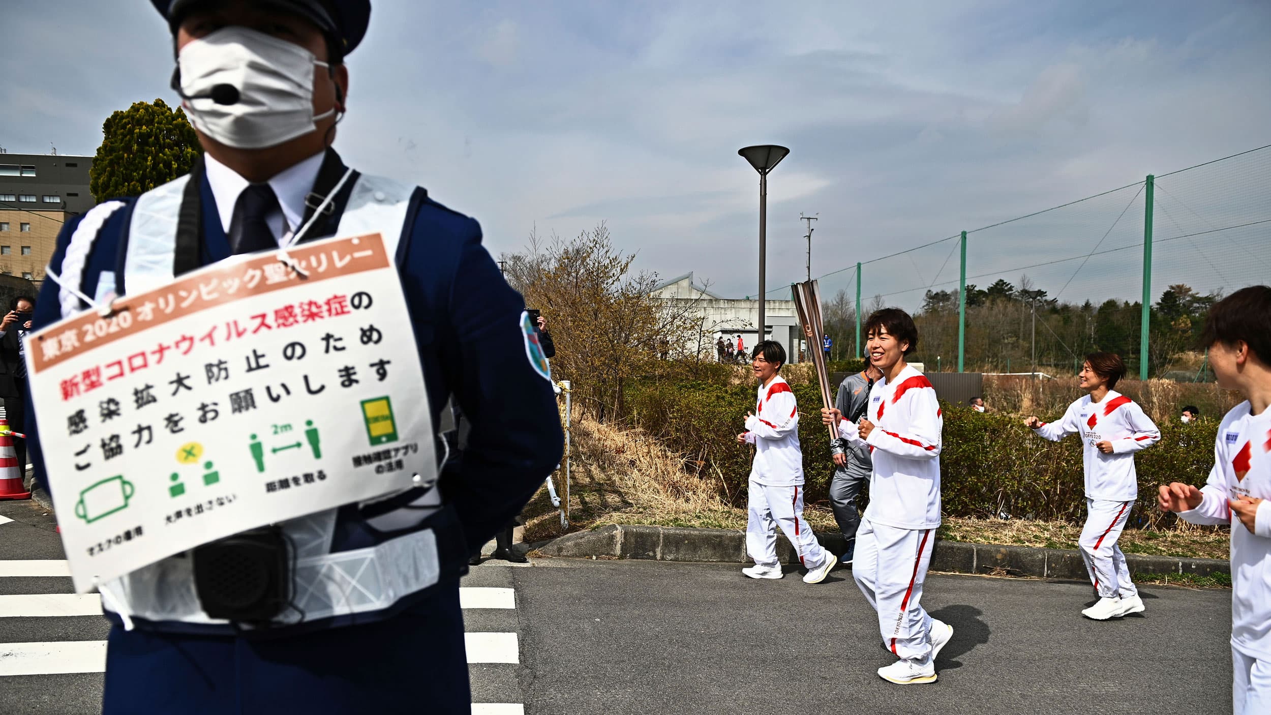 A security official is shown in the nearground with a sign listing COVID-19 safety precautions with several people in roadway behind him and one woman carrying the golden Olympic torch.