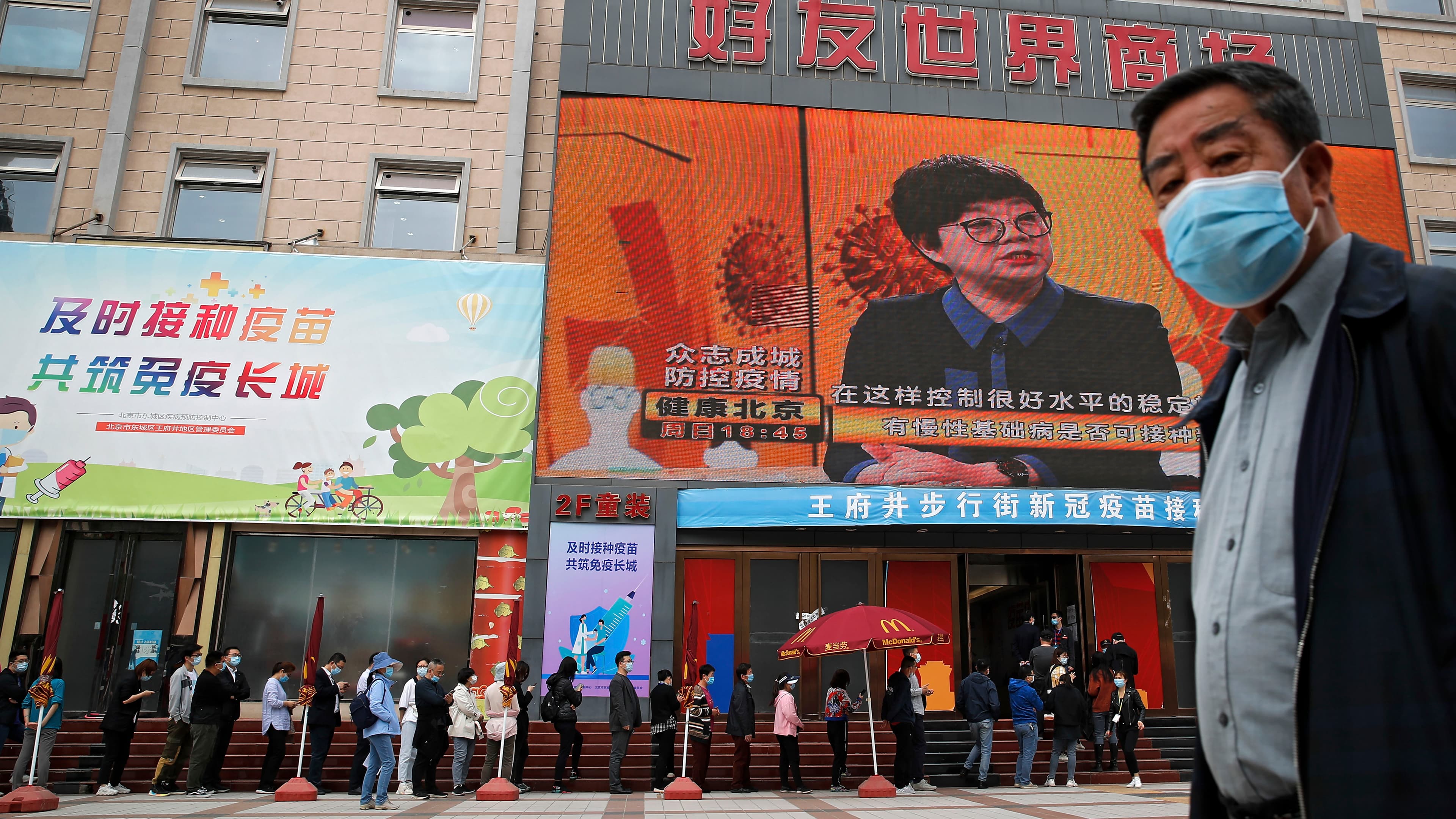 A masked man in a suit stands in front of a line of people waiting for a COVID-19 vaccine.