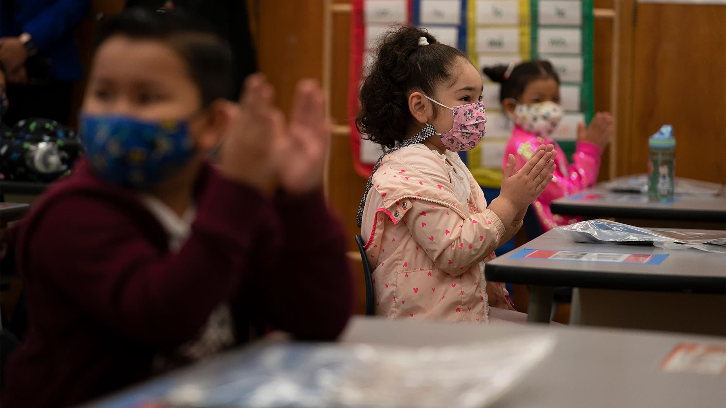 Children in pink and maroon jackets applaud in their classroom