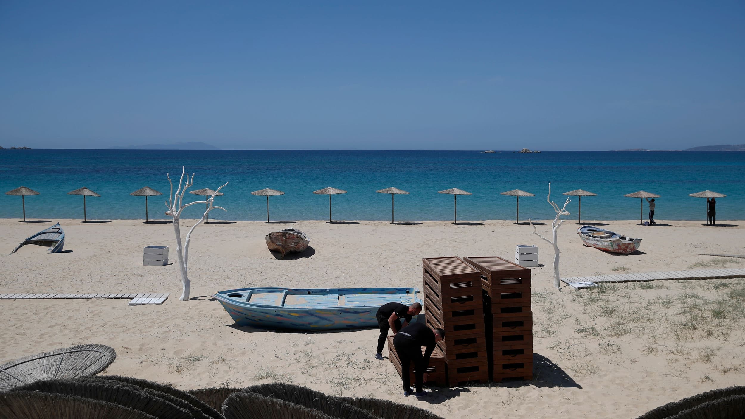 A row of umbrells are shown standing upright in the sand with the blue Aegean sea in the distance.