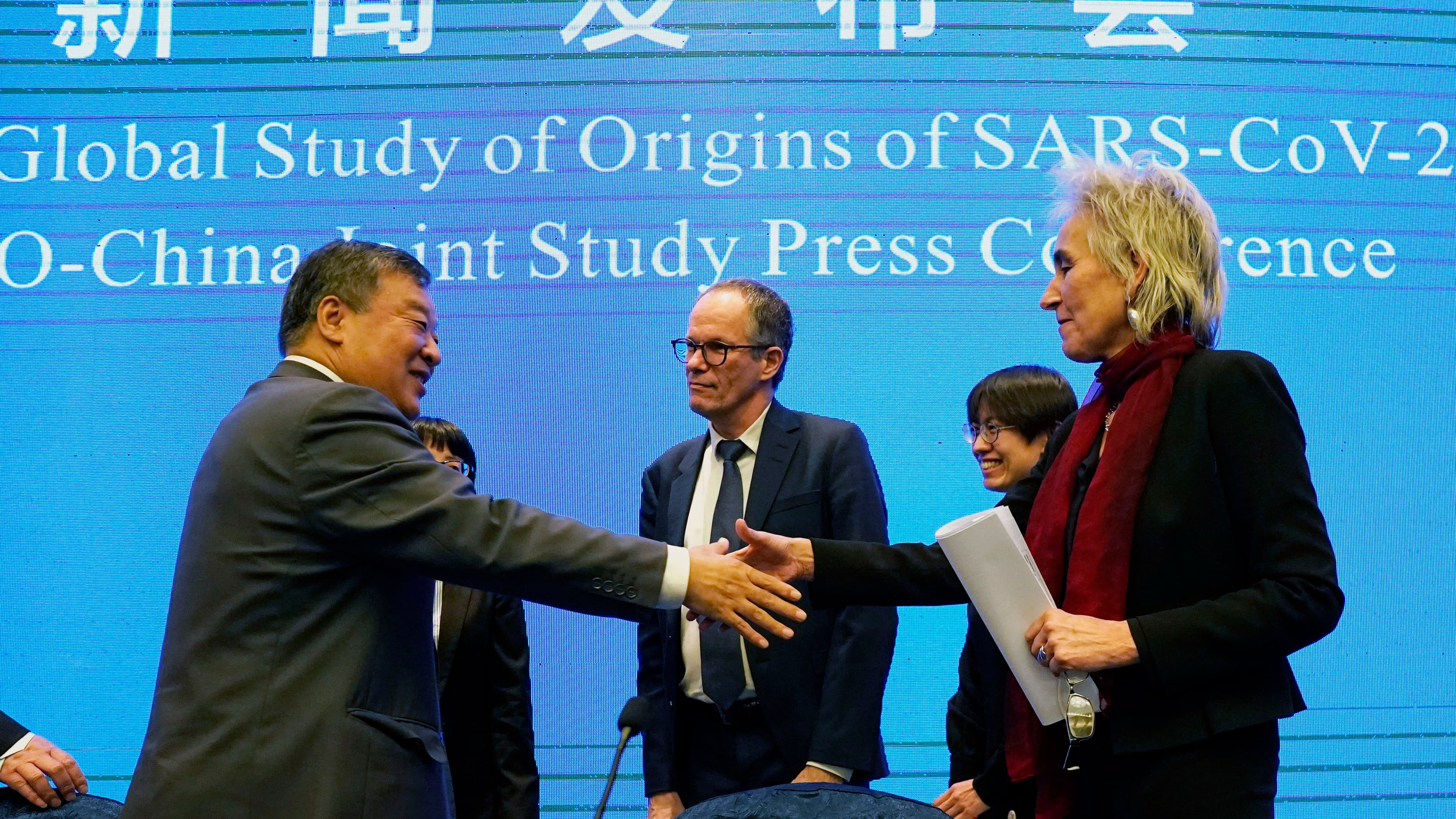 Marion Koopmans, right, and Peter Ben Embarek, center, of the World Health Organization team say farewell through a handshake with a blue banner in the background