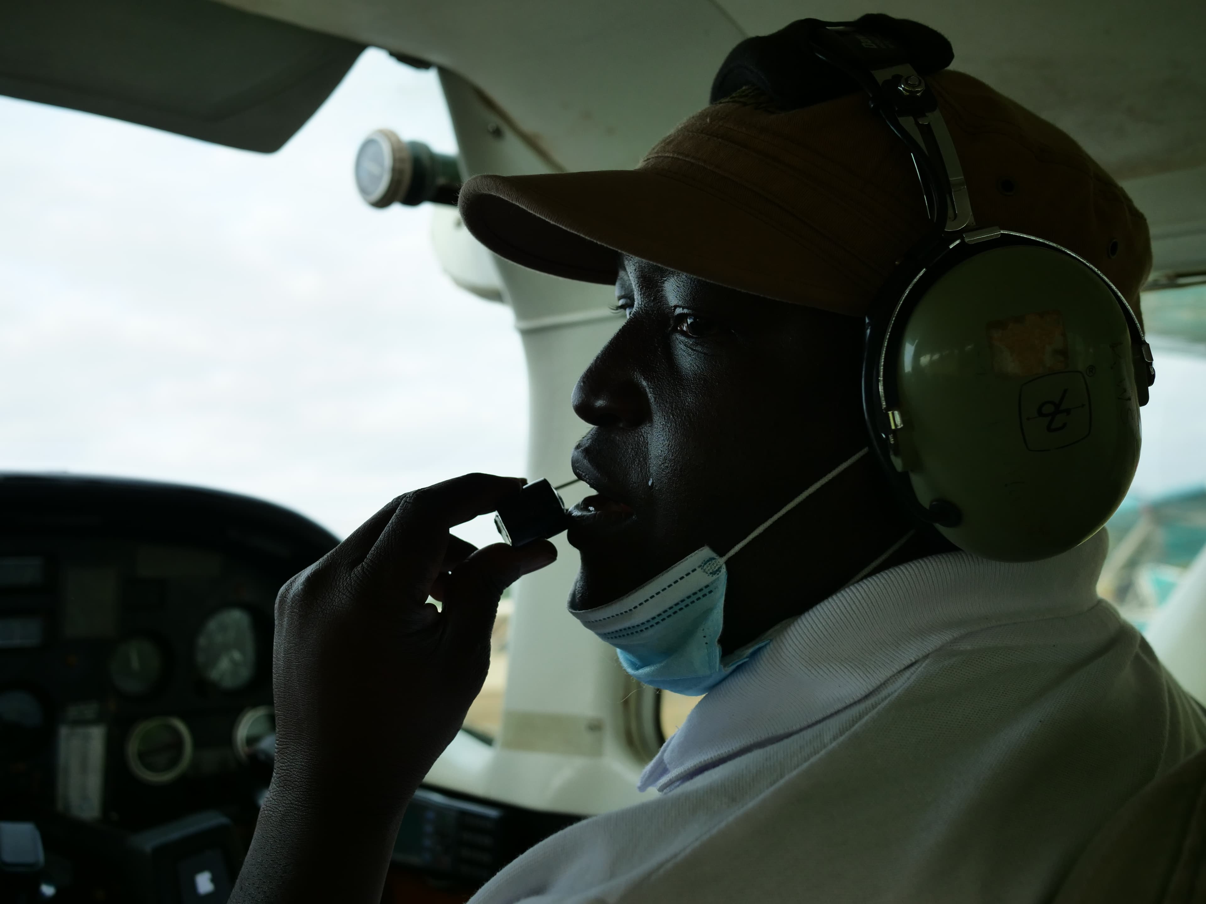 Man in a plane cockpit wearing a hat and headphones speaking into an audio transmitter