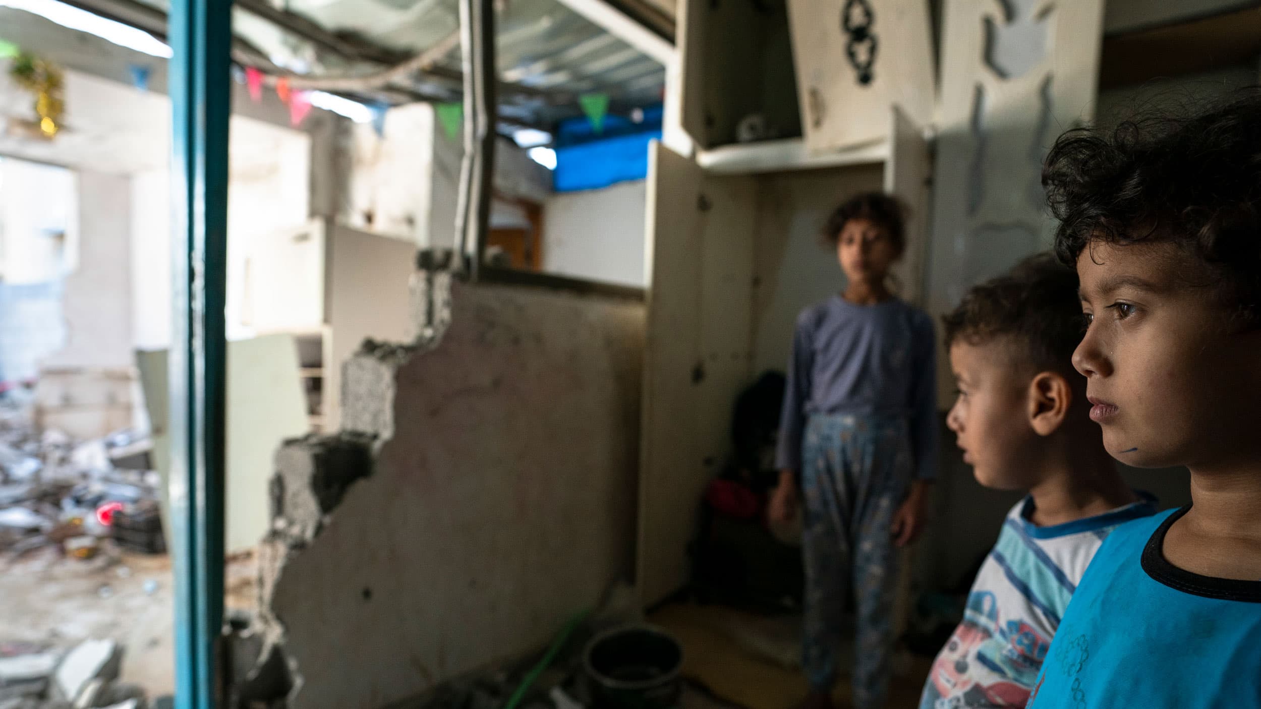Three young children are shown looking off to a room damaged by rocket attack.