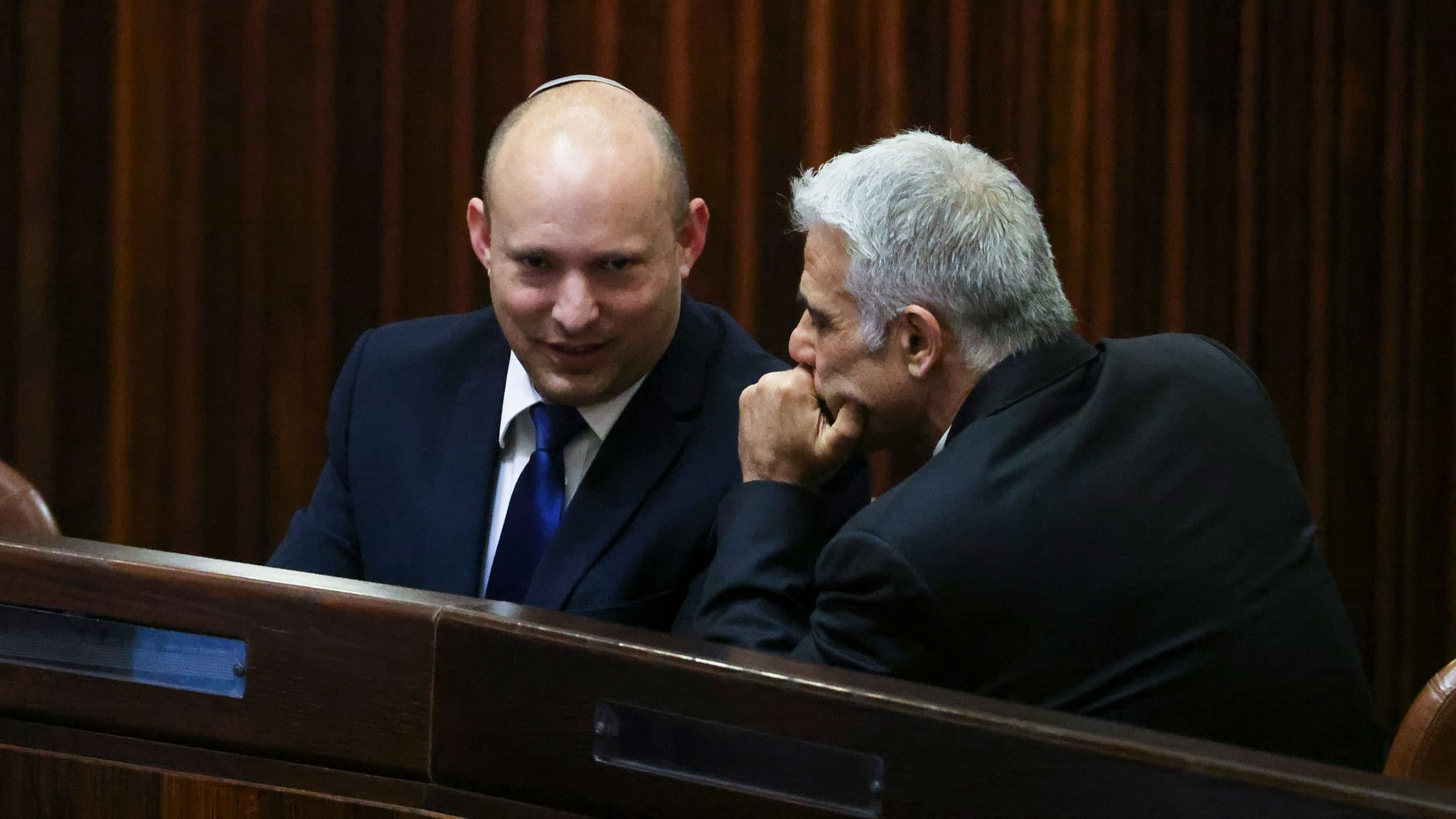 Yamina party leader Naftali Bennett wearing a dark suit and sitting while speaking with Yesh Atid party leader Yair Lapid on his left.