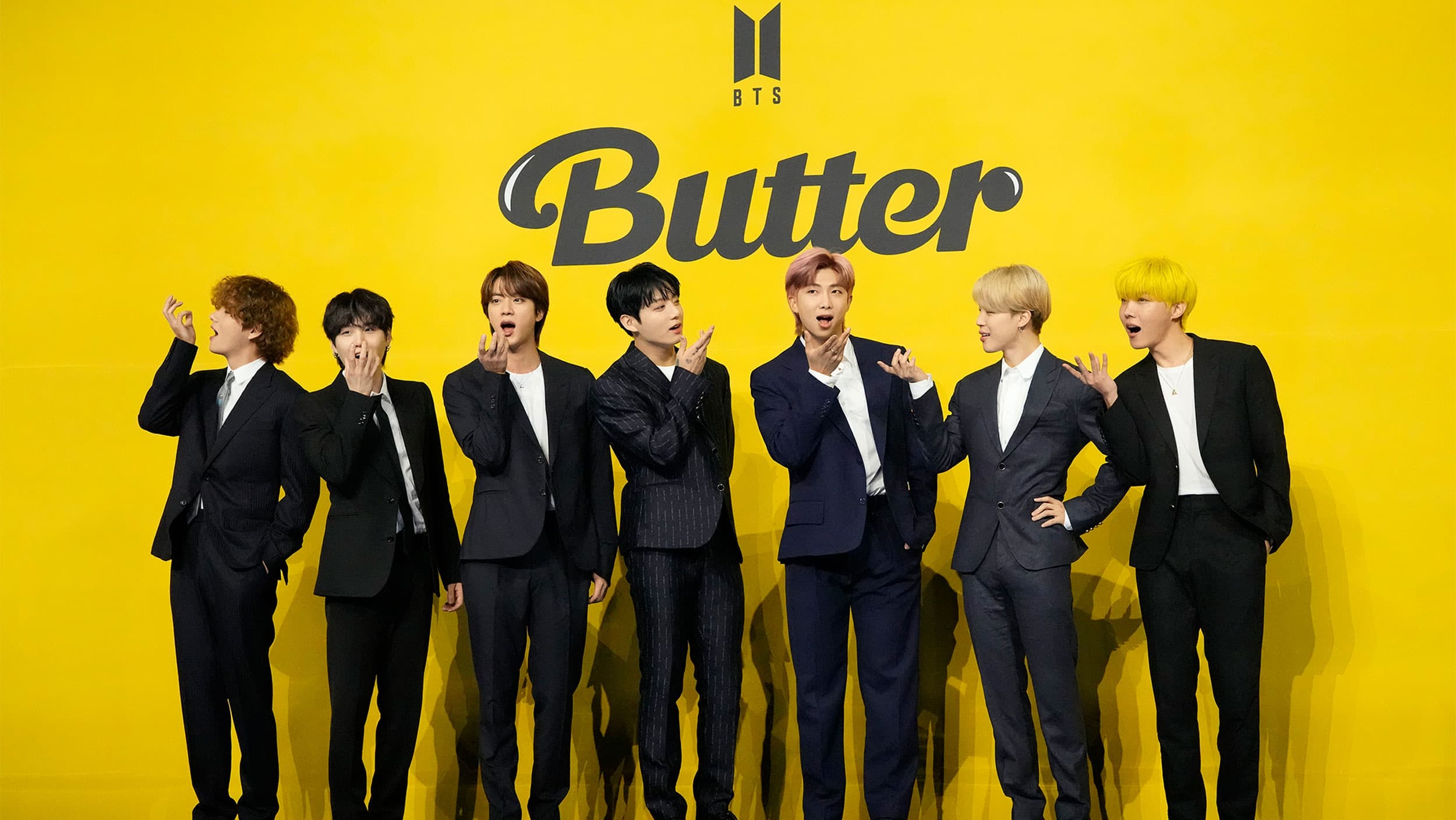Seven members of K-pop band BTS stand wearing black suits in front of a yellow background that says 
