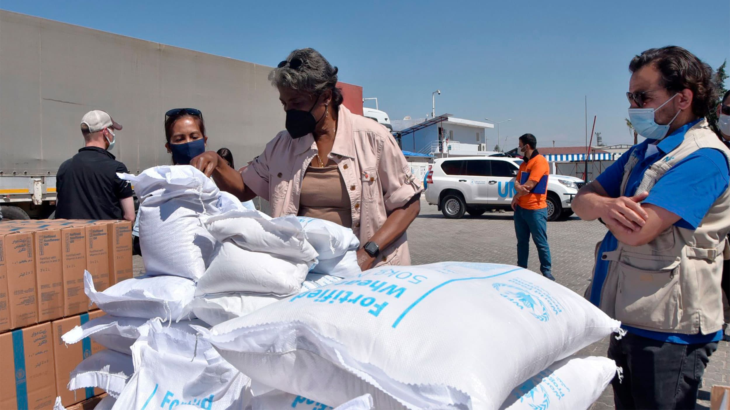 People wearing masks stand in front of white bags of food aid