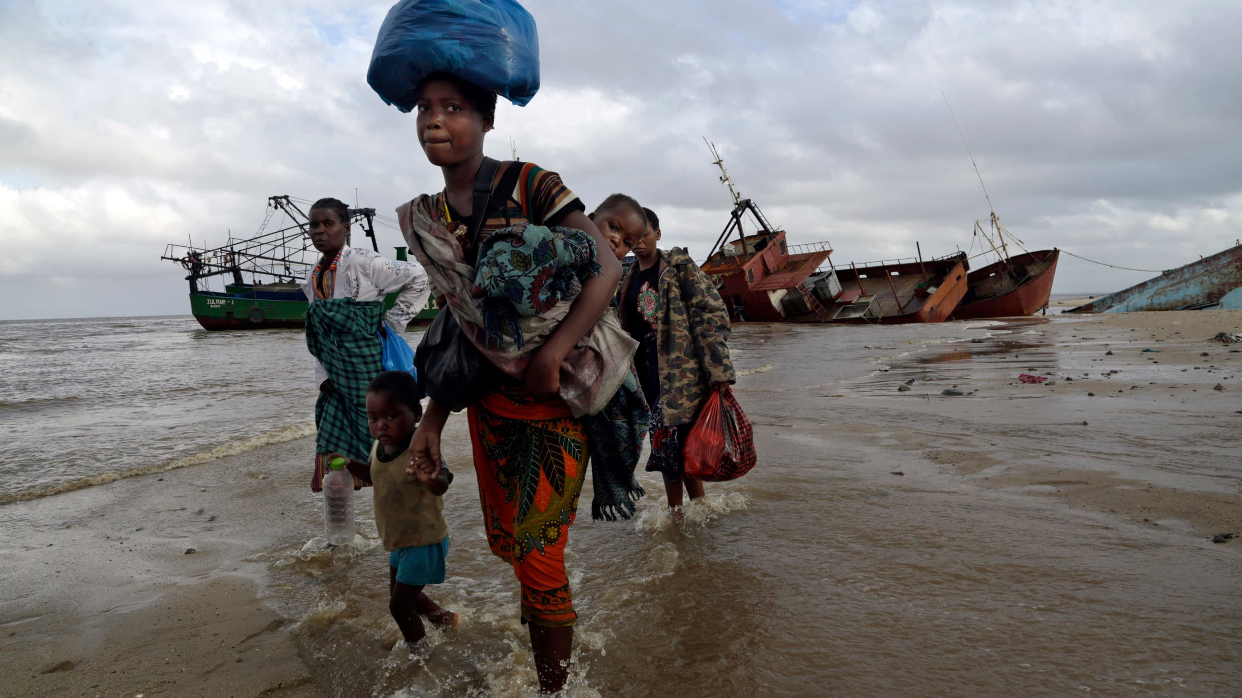 A small group of people are shown walking through coastal waters in Mozambique with large ships in the distance.