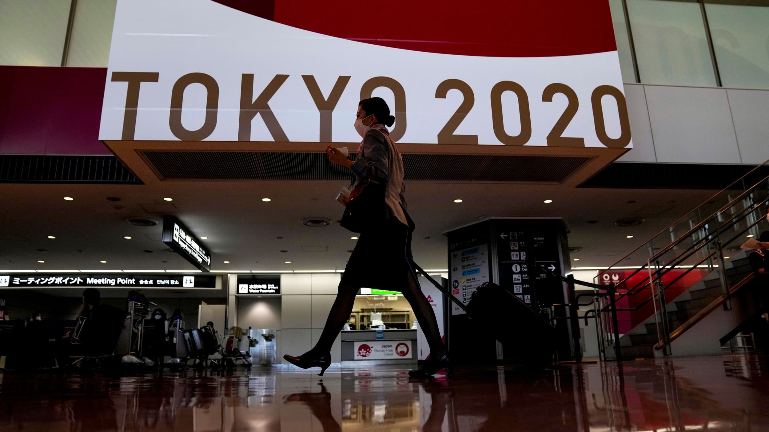 A woman is shown walking and pulling a rollaboard in an airport terminal with the Tokyo Olympics 2020 sign in the background.