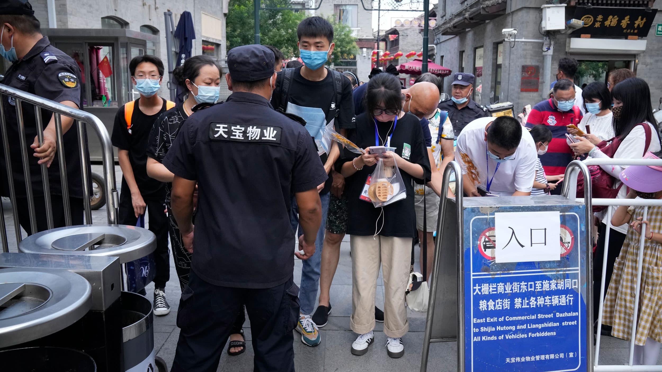 A crowd of people are shown standing at the temporary gated entrance where a security official is checking health codes.